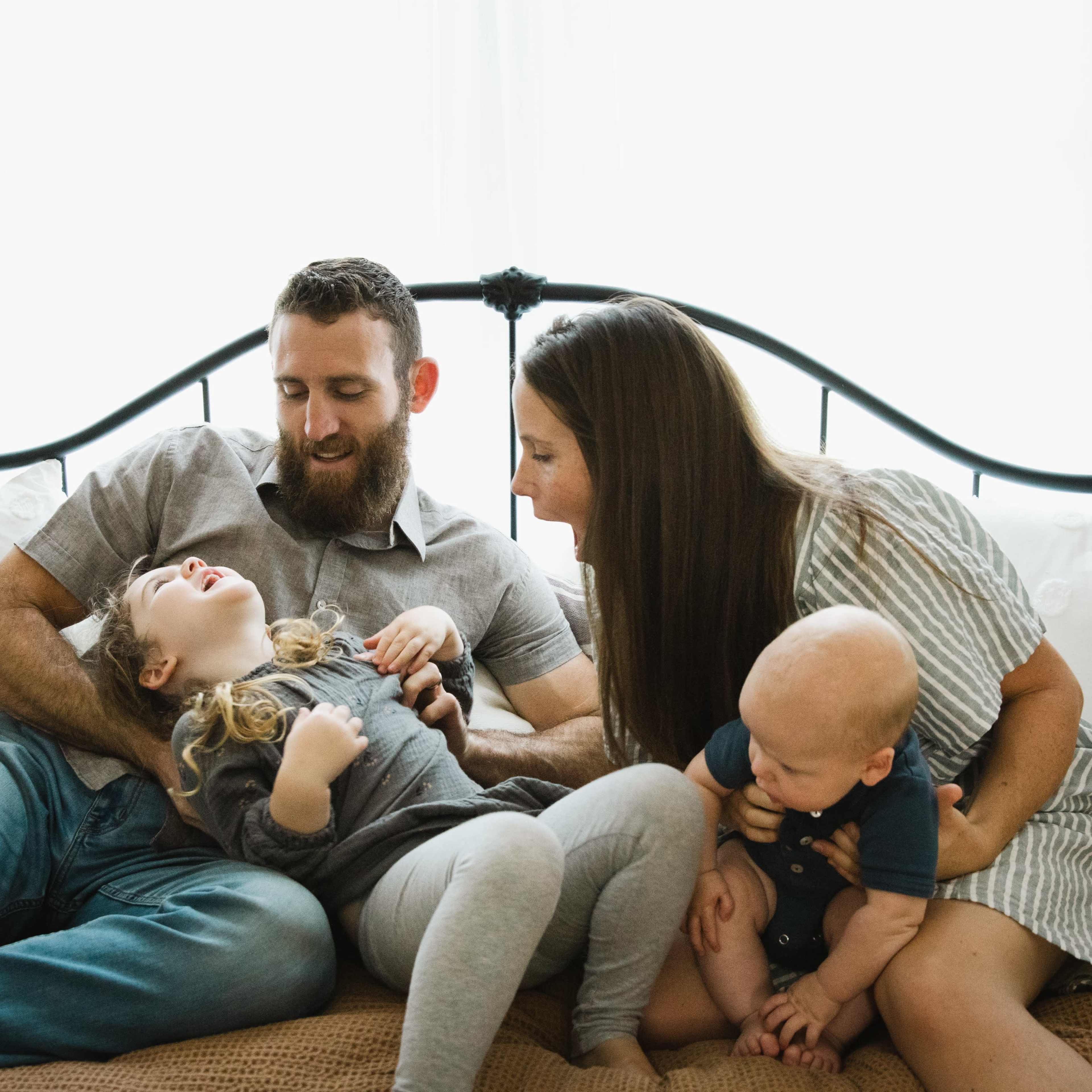 A father and mother sit on a bed with their two young children, engaging in playful interaction.