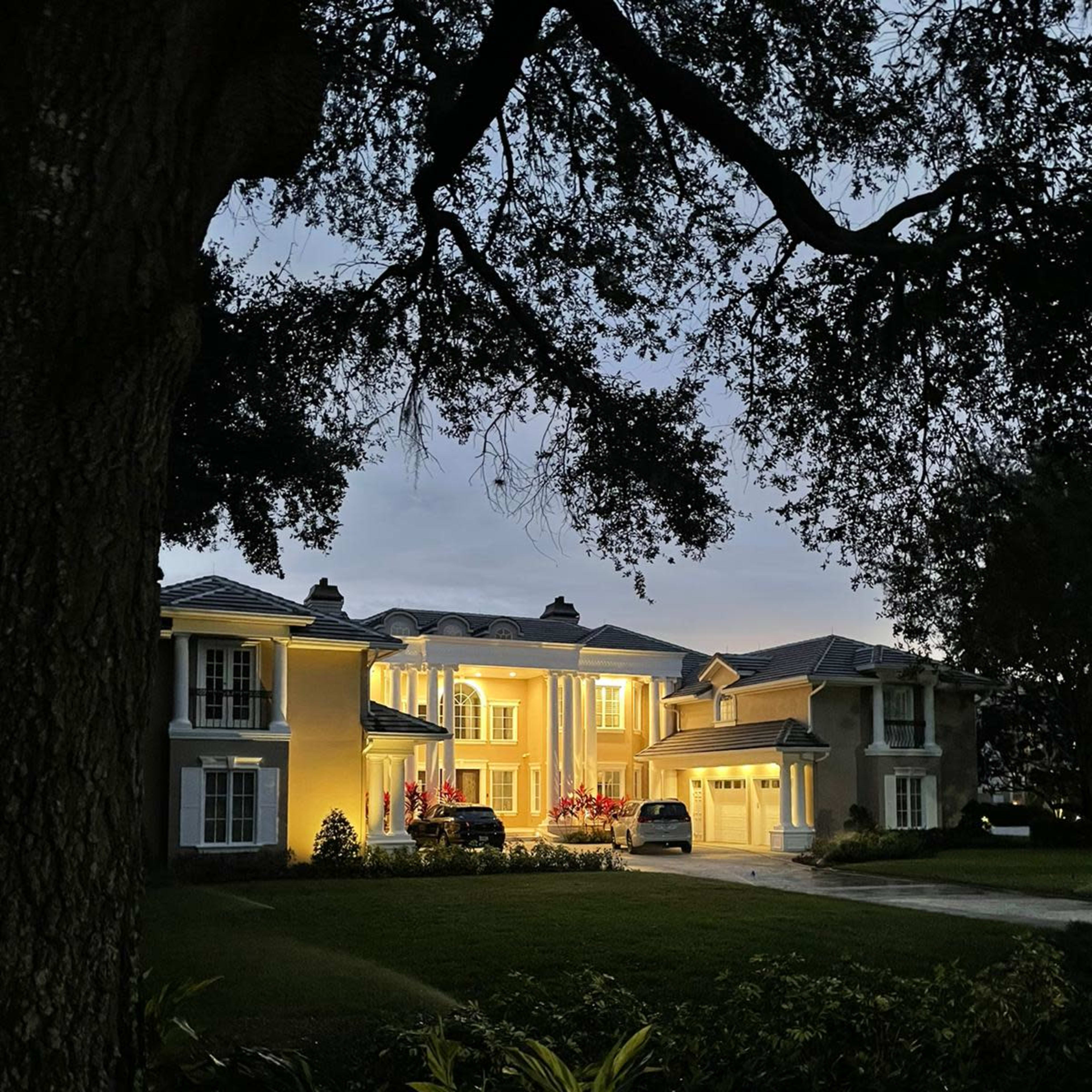 A well-lit, two-story house with a manicured lawn is shown during twilight, surrounded by trees and shrubs.