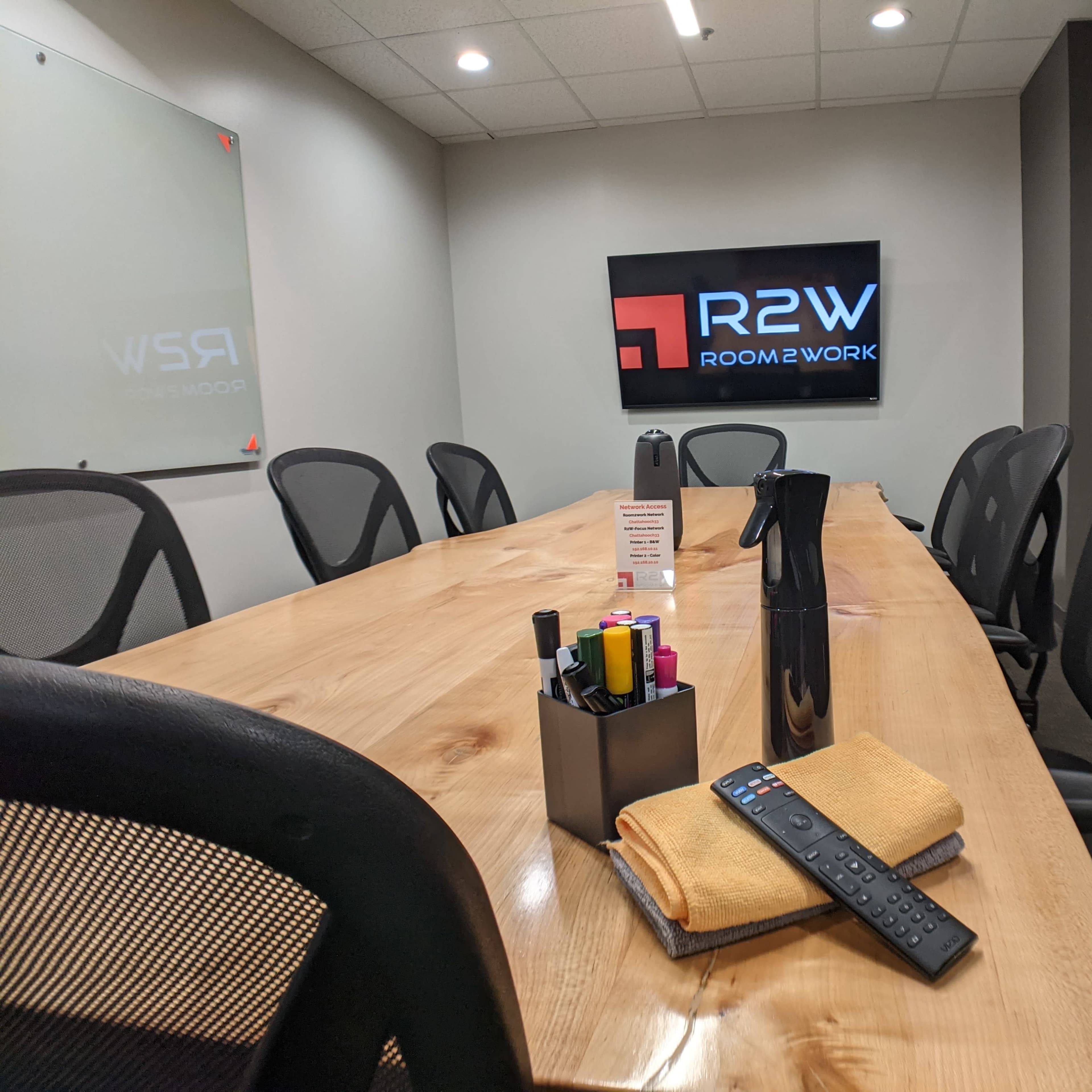 A conference room features a long wooden table with office supplies, a remote control, and a television displaying the logo "R2W Room2Work."