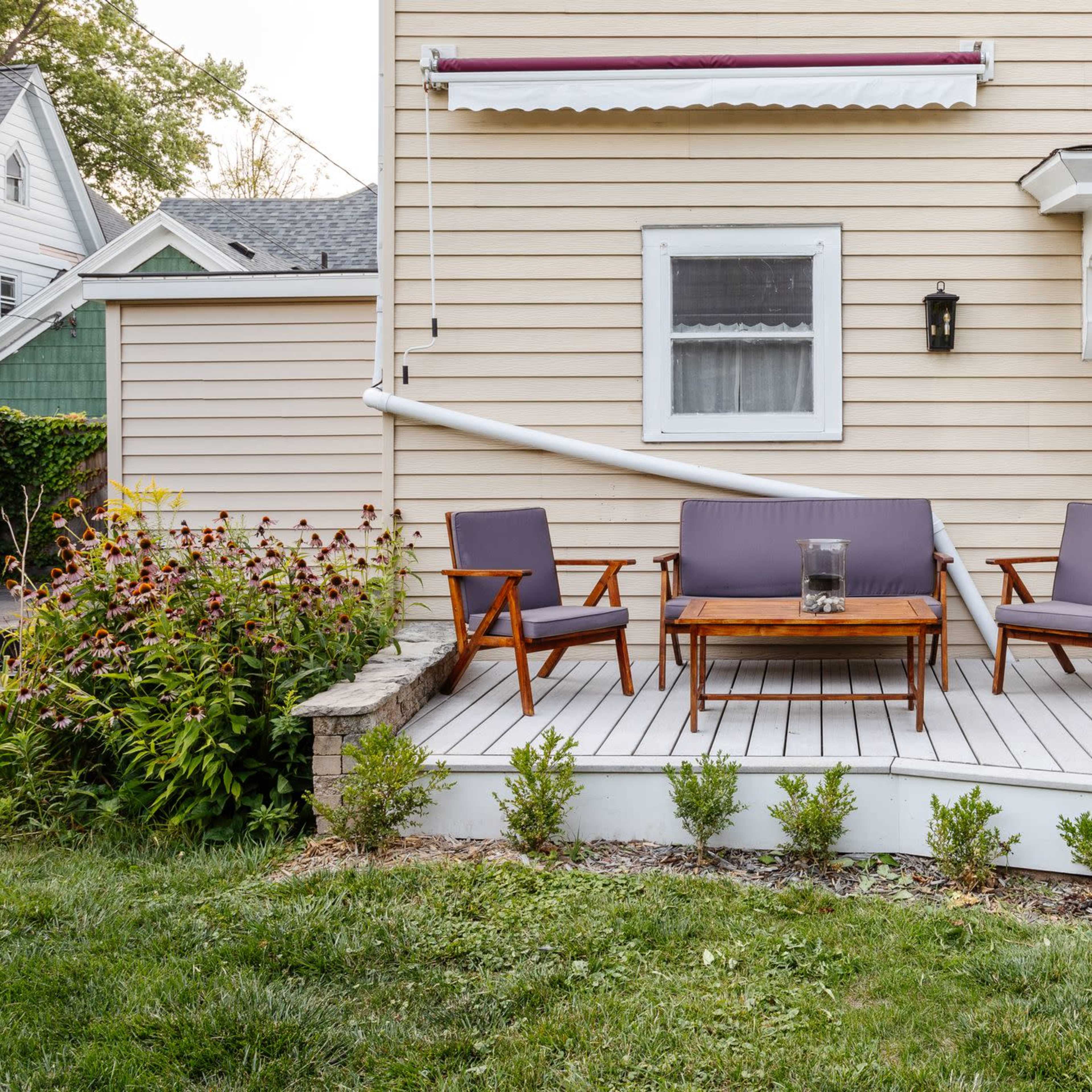A wooden deck features three chairs and a table in front of a house with beige siding and a white door.