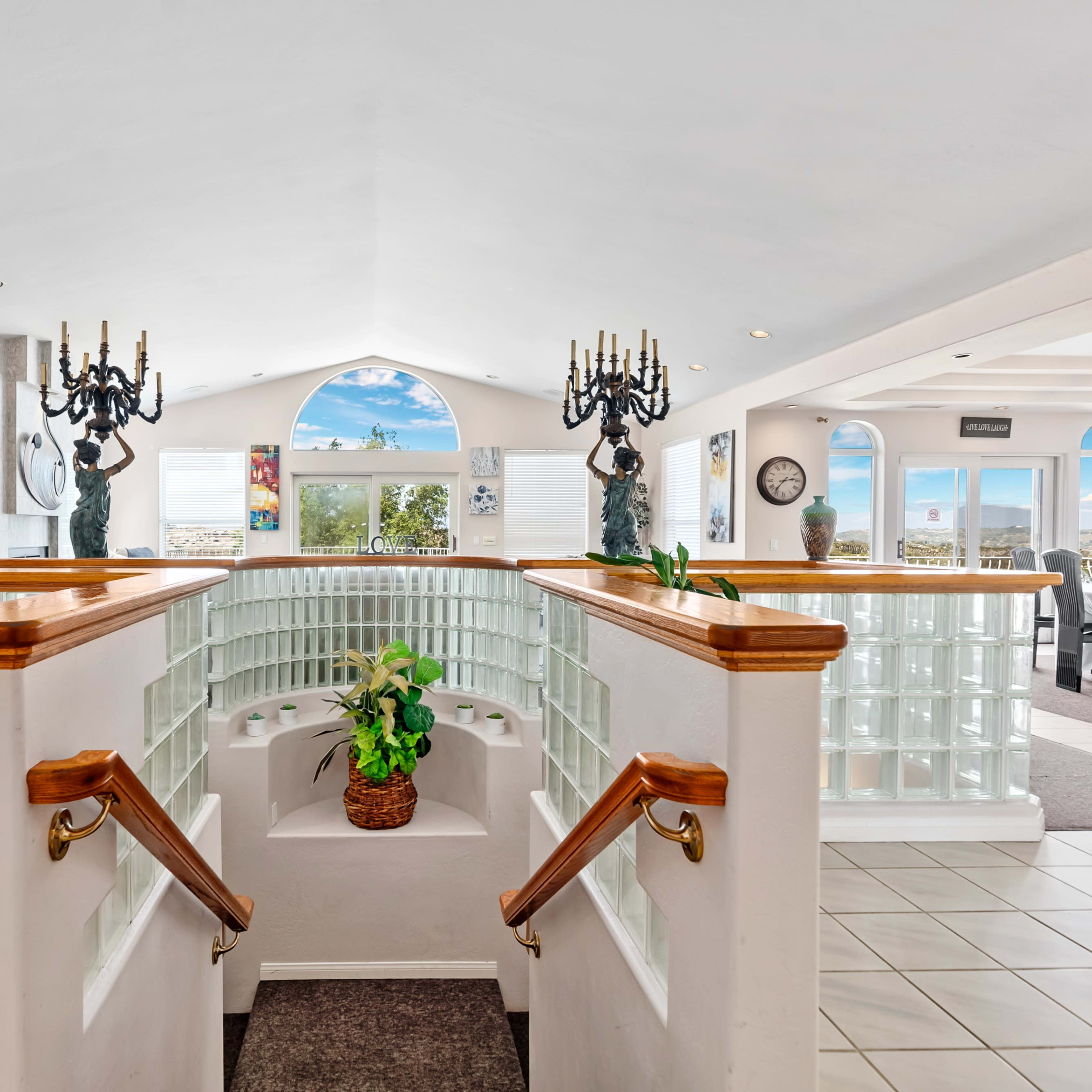 A spacious interior featuring a curved staircase with glass block railings, potted plants, and large windows providing a view of the sky.