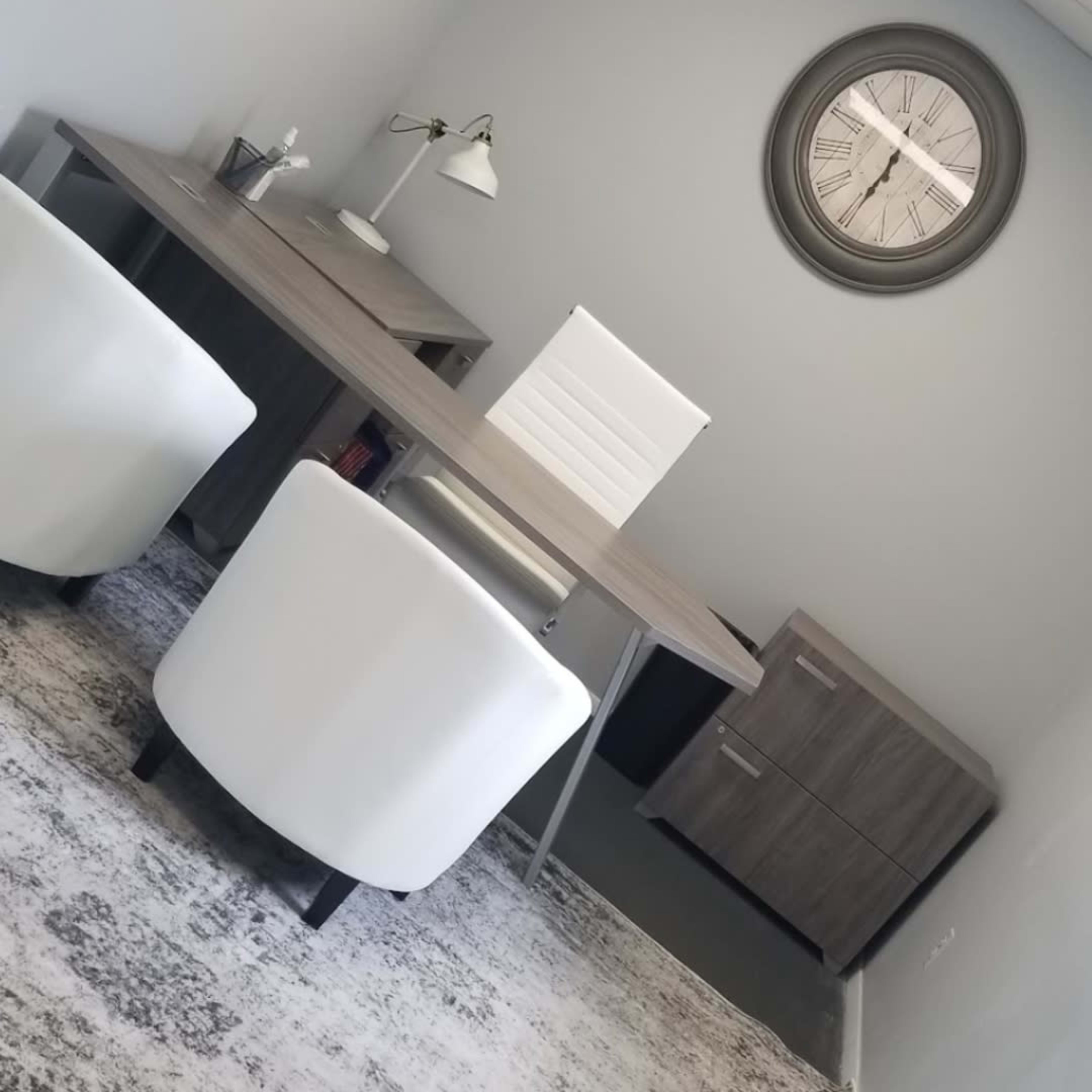 A modern office space featuring a large desk, two white chairs, a wall clock, and a gray area rug.