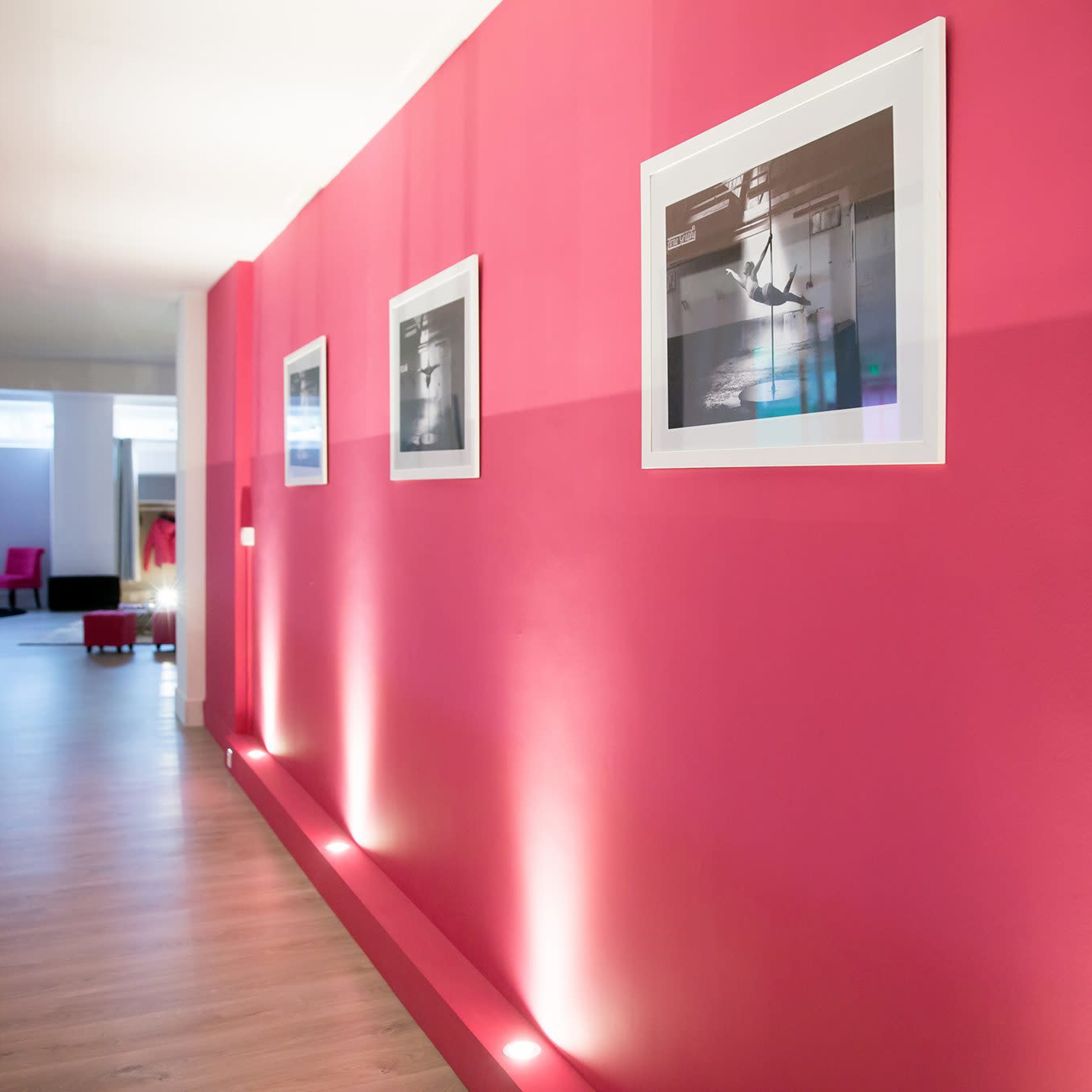 A corridor with pink walls features framed photographs and subtle lighting along the floor.