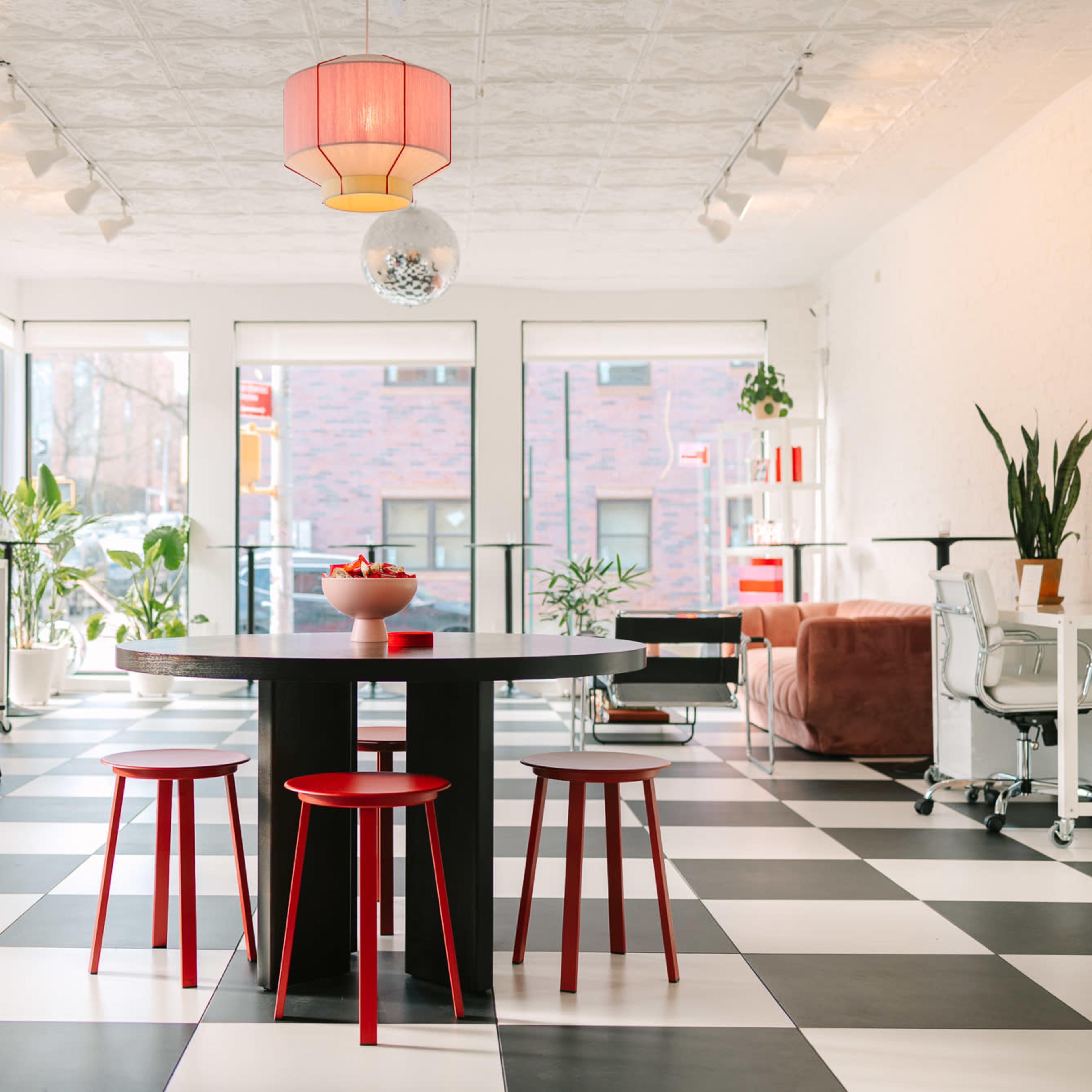 A modern room features a black and white checkered floor, a round table with three red chairs, and large windows filled with plants and natural light.