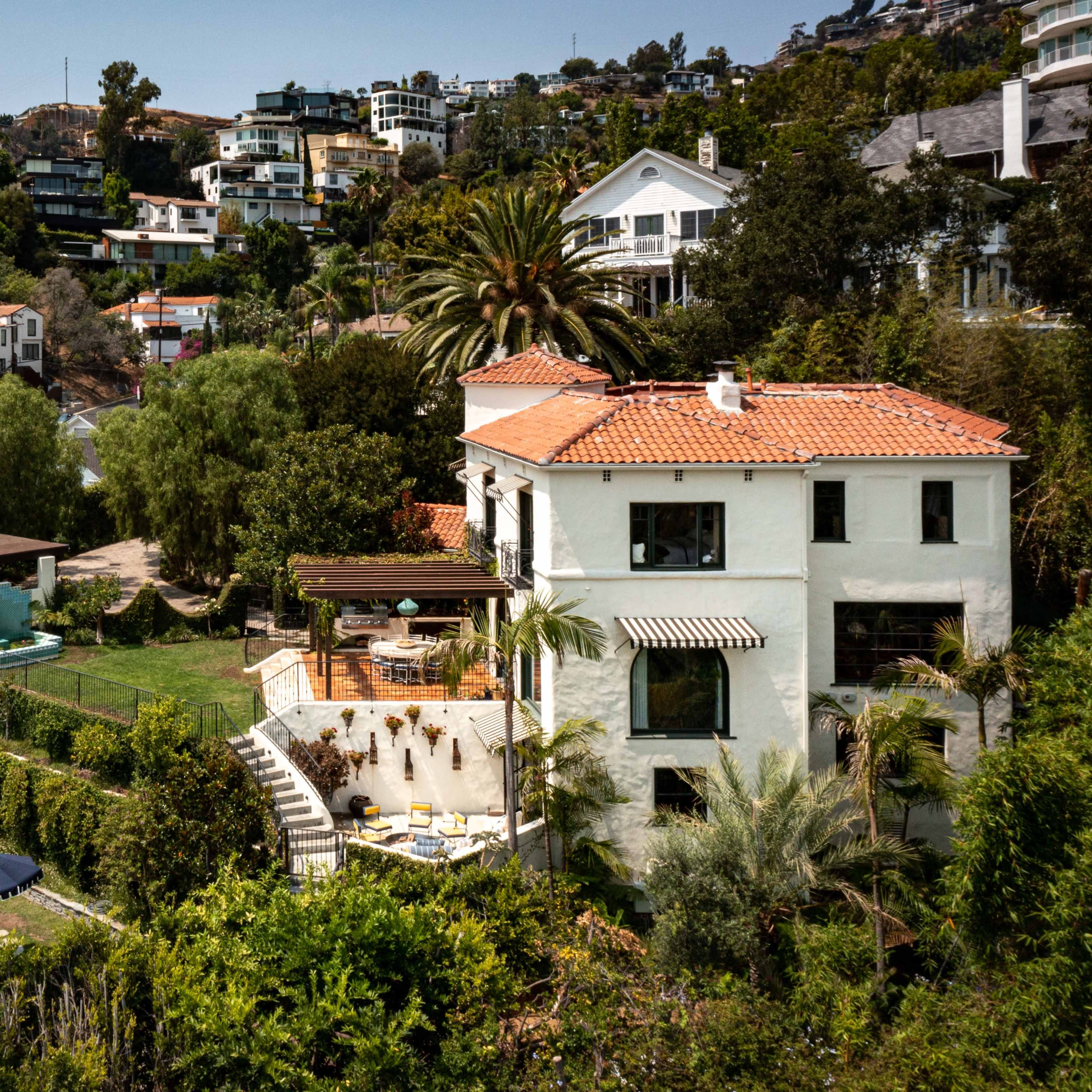 A multi-level white house with a red tile roof surrounded by greenery, a swimming pool, and several outdoor seating areas, situated in a hilly residential area.