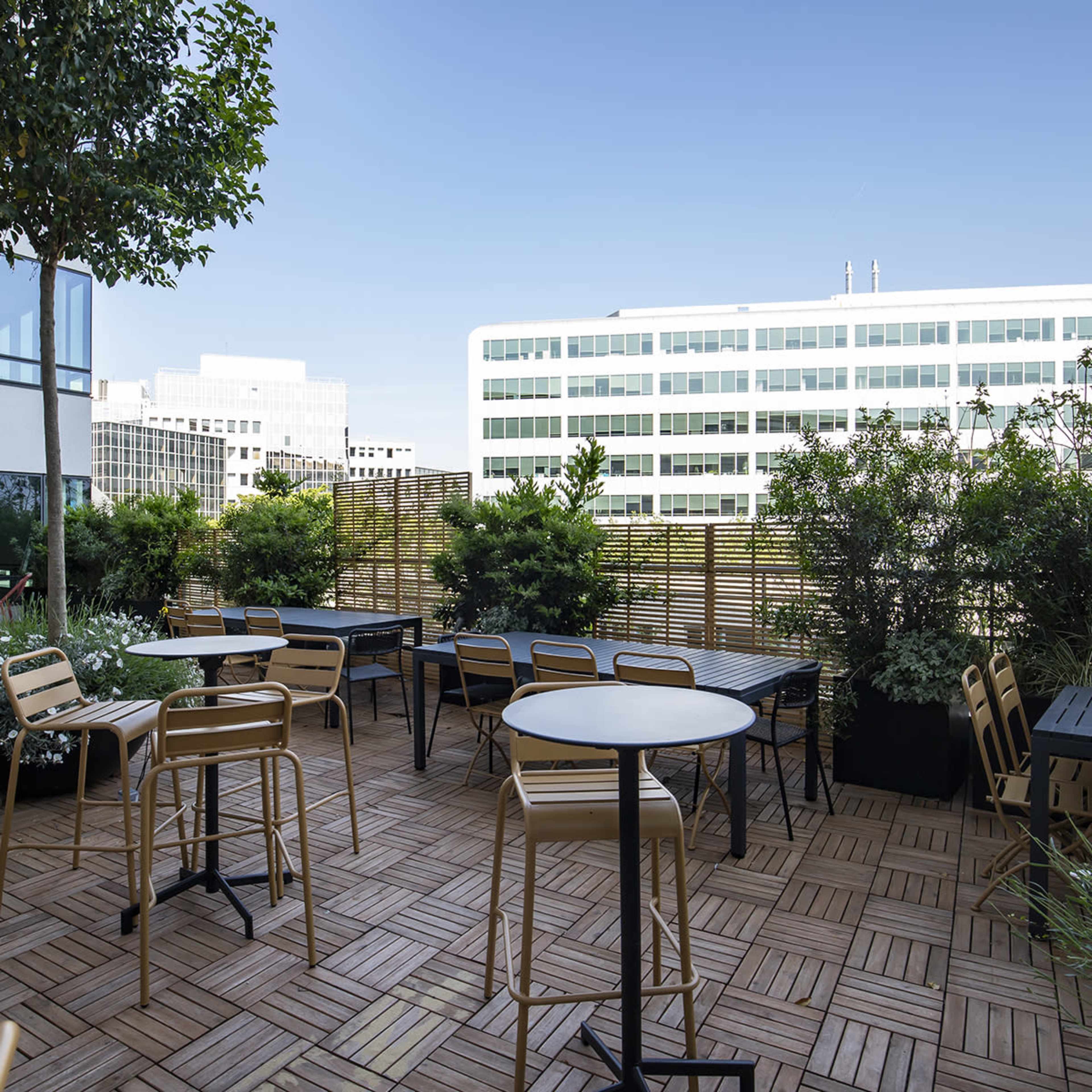 A rooftop patio featuring several tables and chairs surrounded by greenery and a modern building backdrop.
