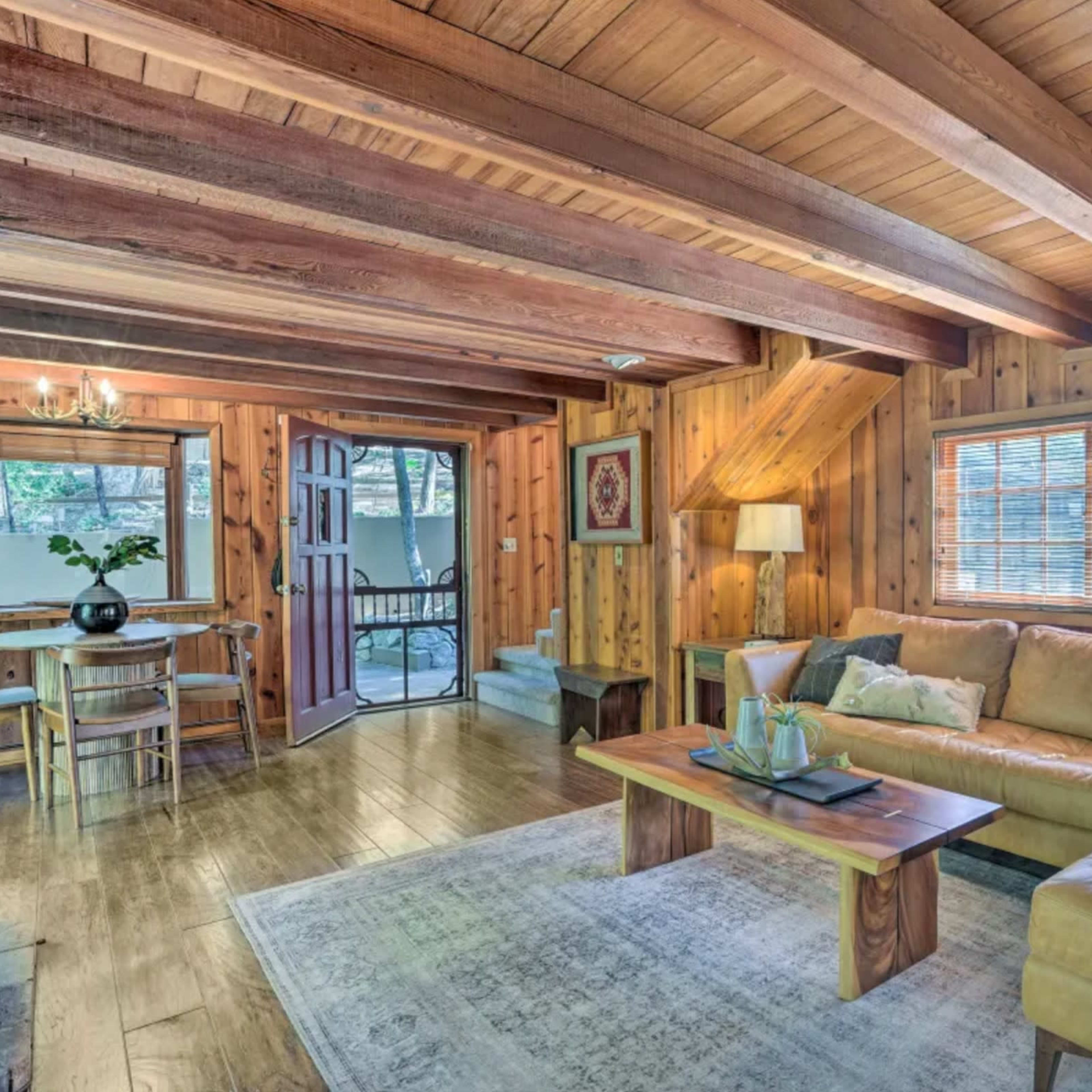 The room features wooden beams and walls, a tan sofa, a coffee table, a dining area, and a door leading outside.