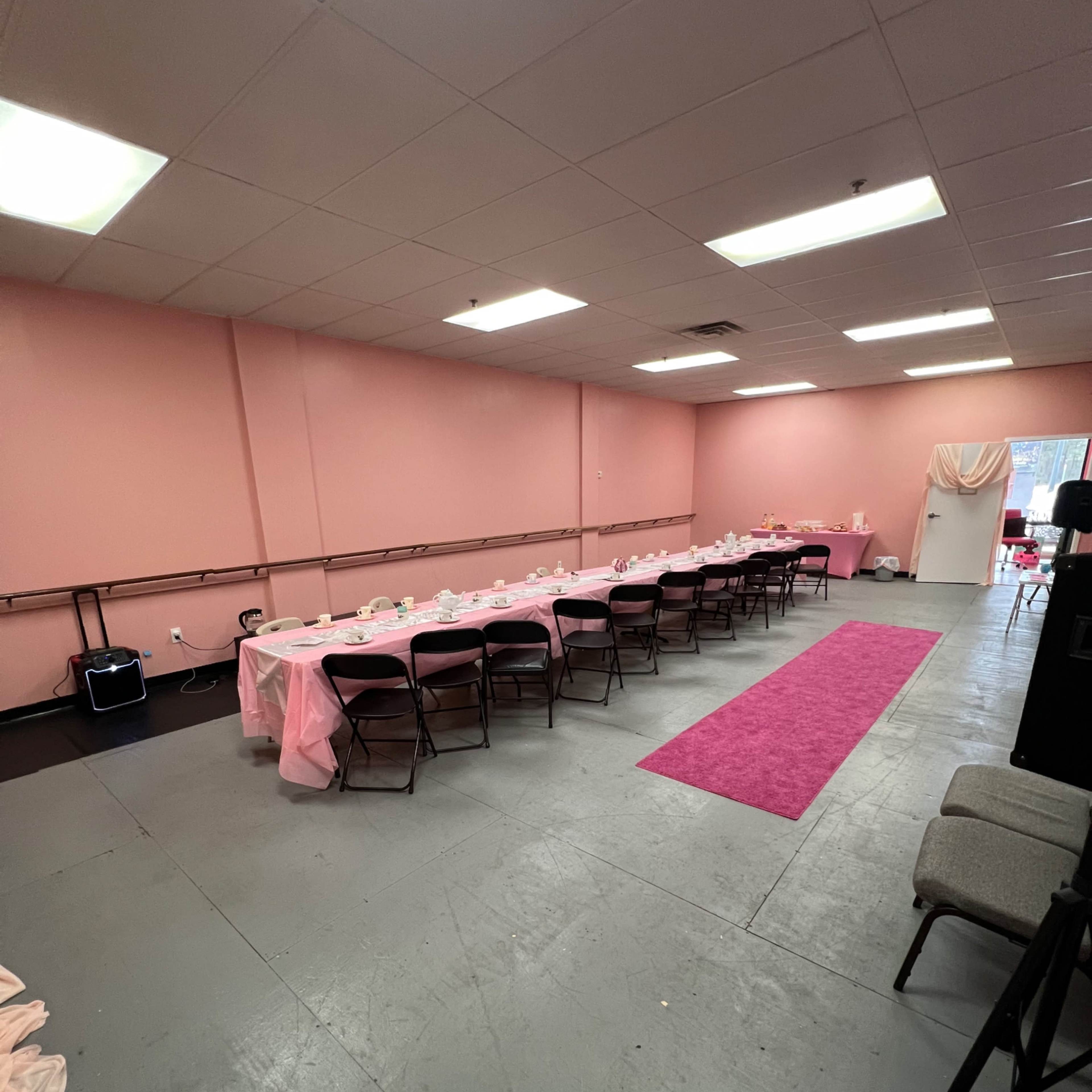 A pink-themed event space features a long table set with chairs, centered on a pink carpet, against a backdrop of pink walls.