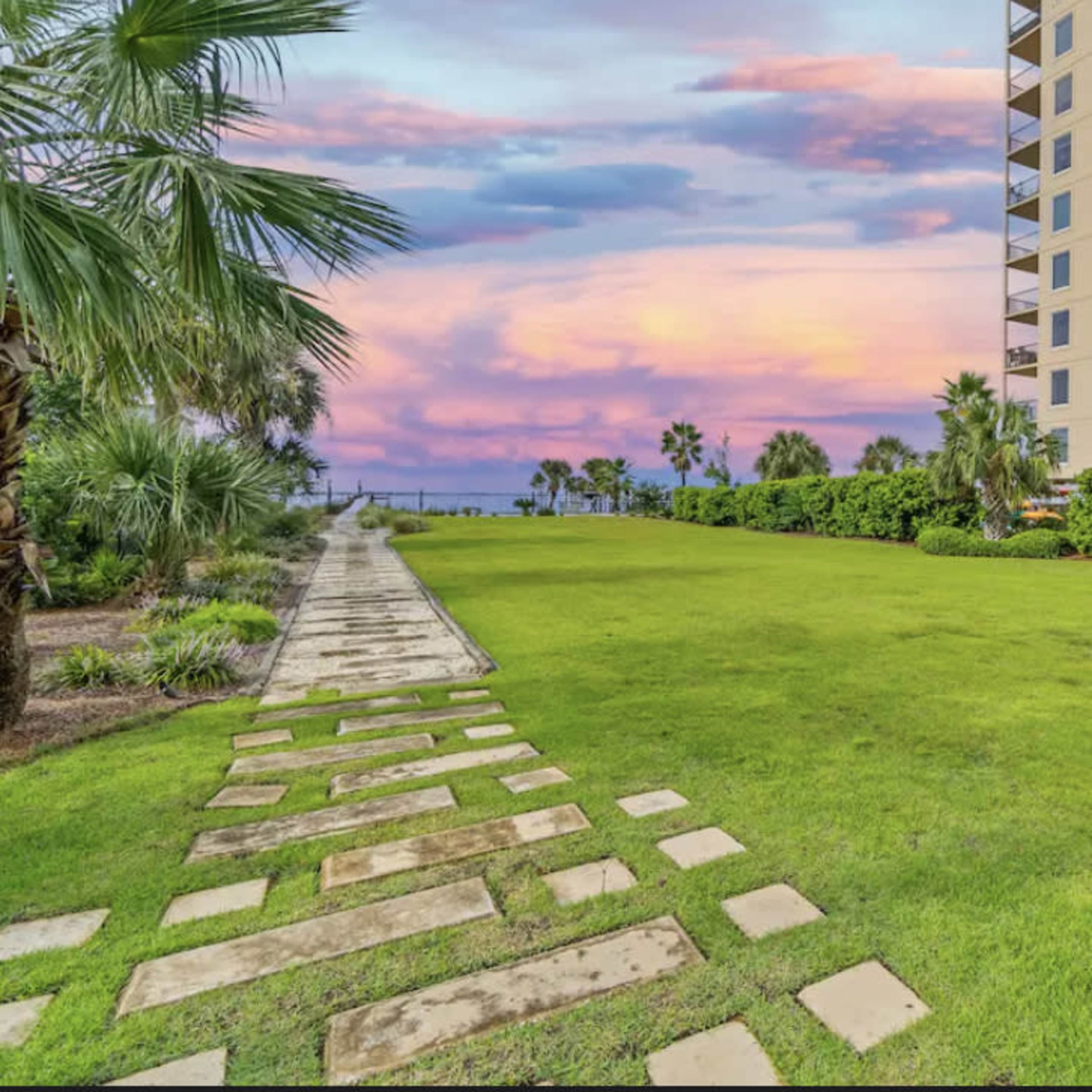 A stone pathway leads through a grassy area bordered by palm trees, with a colorful sunset reflected in the sky above the ocean in the background.