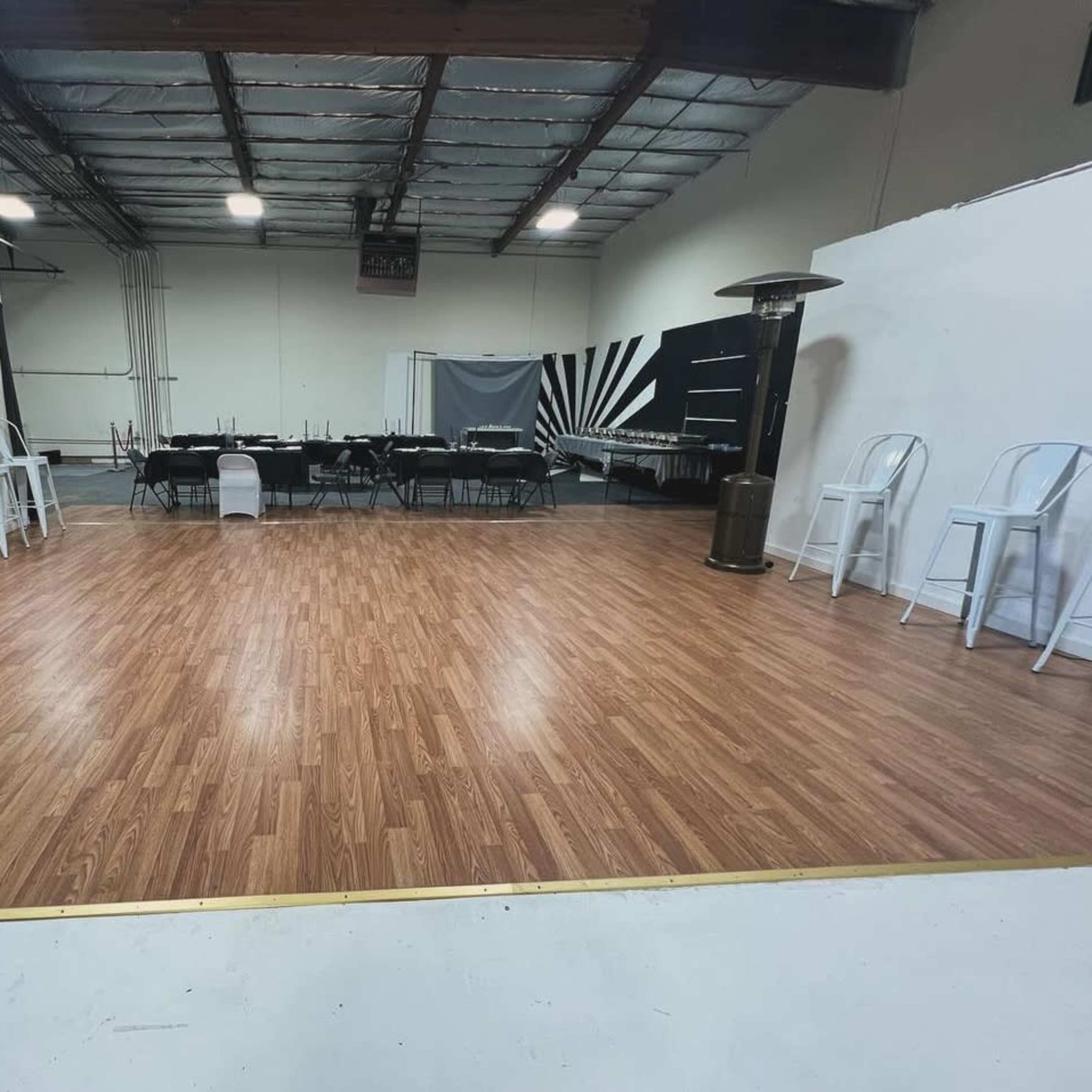 A spacious interior room with a polished wood floor, a long black table set up for seating, and a few chairs arranged along the walls.
