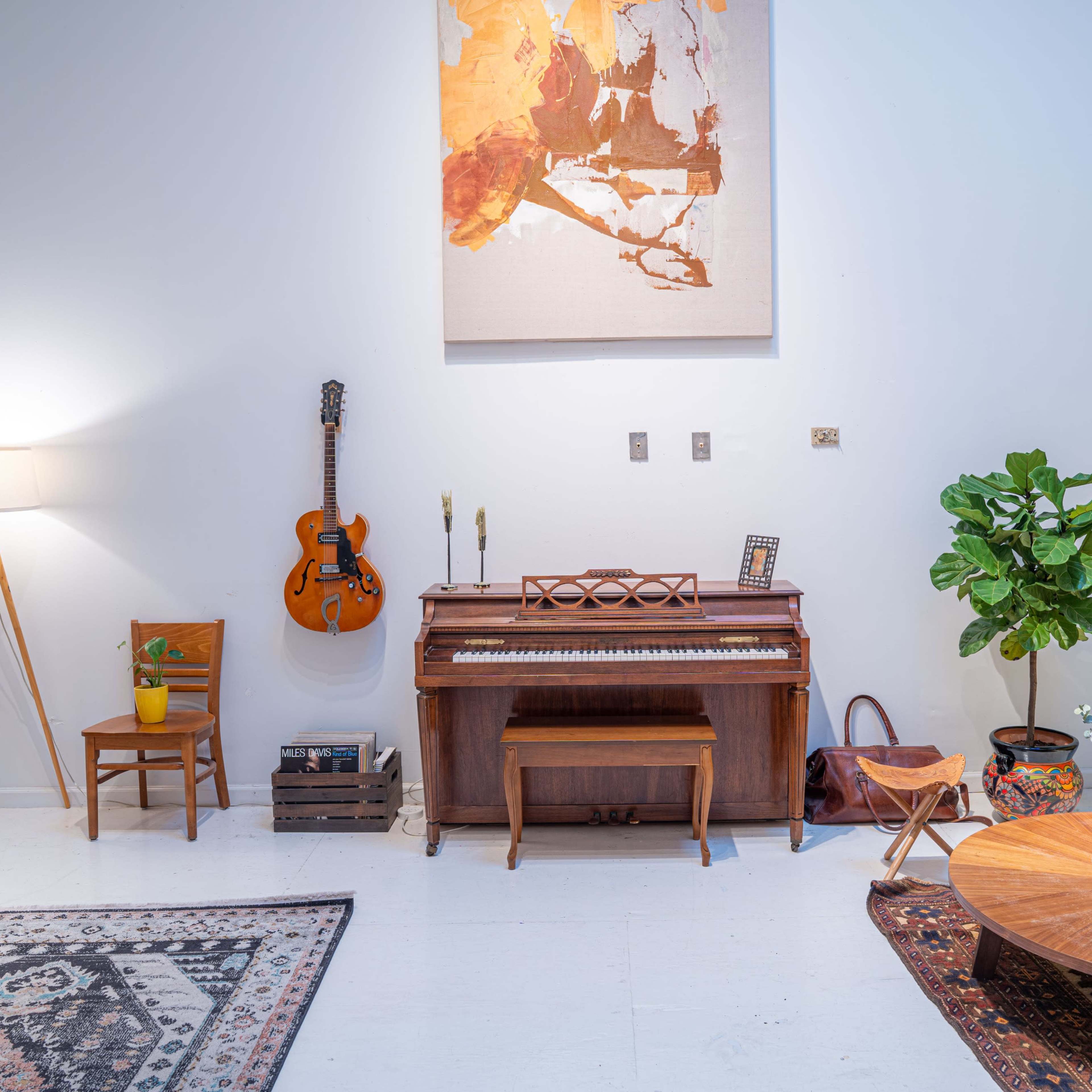 A cozy living room with a piano, a guitar hanging on the wall, a round coffee table, plants, and a light-colored sofa.