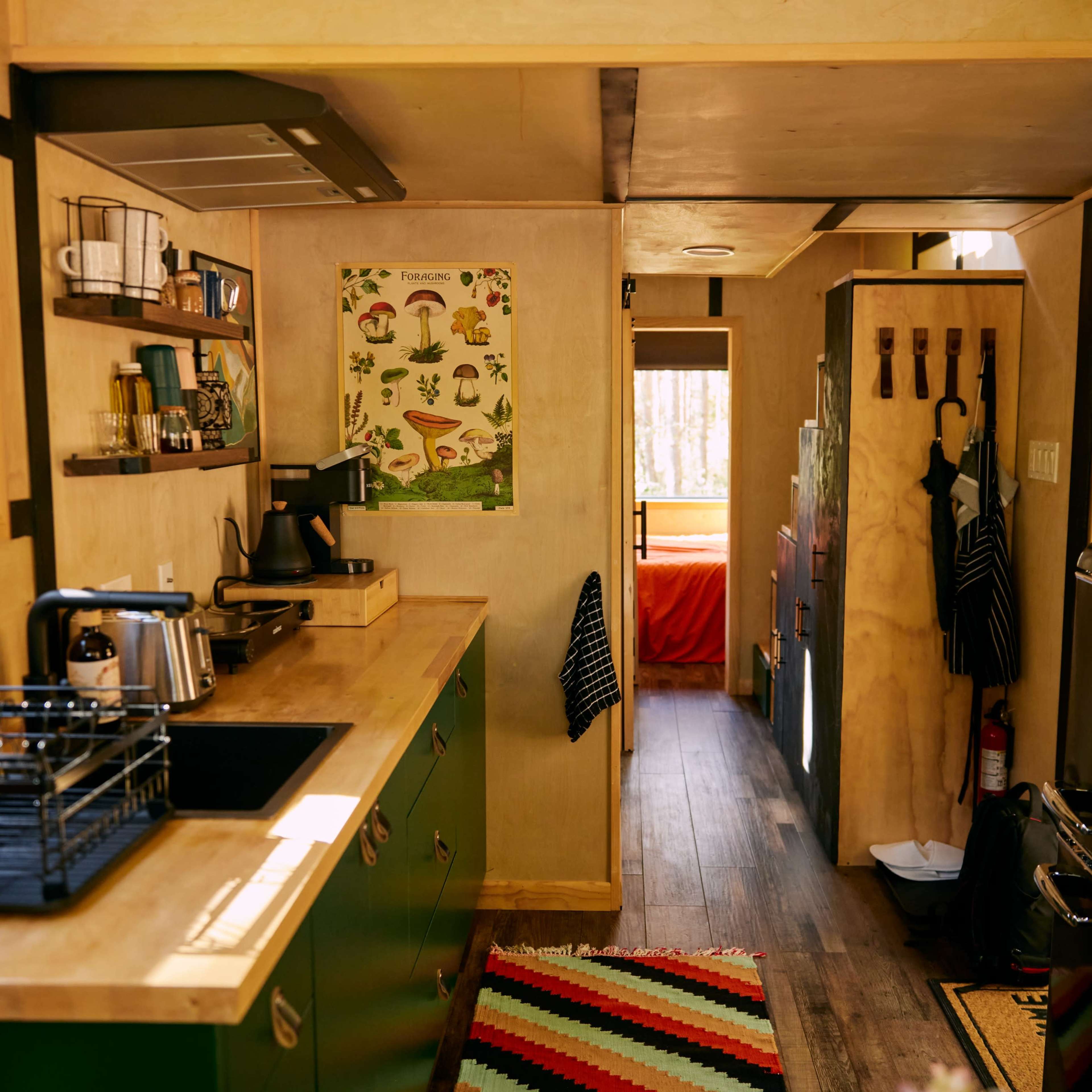 The interior of a compact kitchen in a tiny house, featuring a wooden countertop, green cabinetry, and a view down a hallway toward a bedroom area.