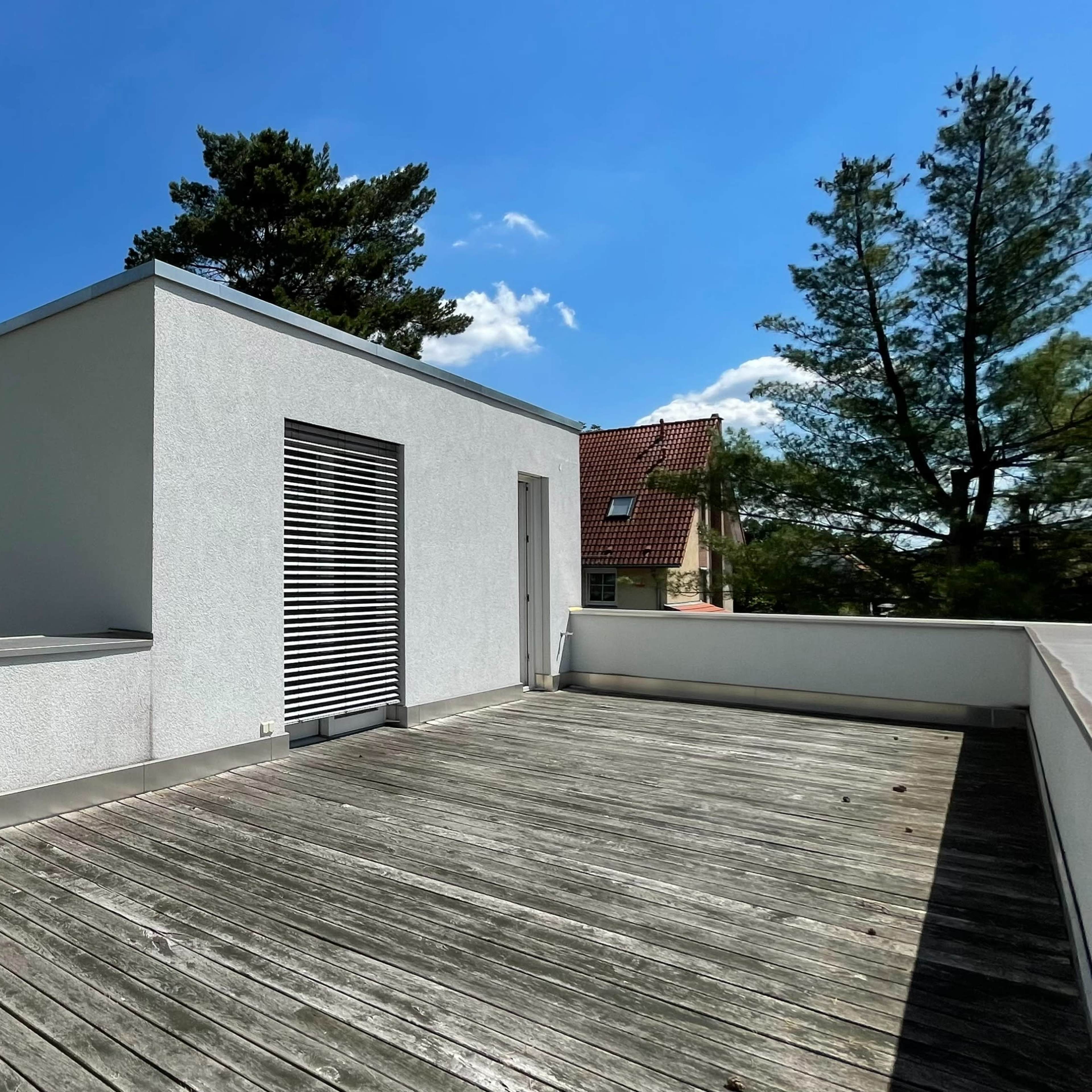 A flat rooftop terrace features wooden decking and a sleek, white-painted structure with a horizontal slatted door, set against a clear blue sky.