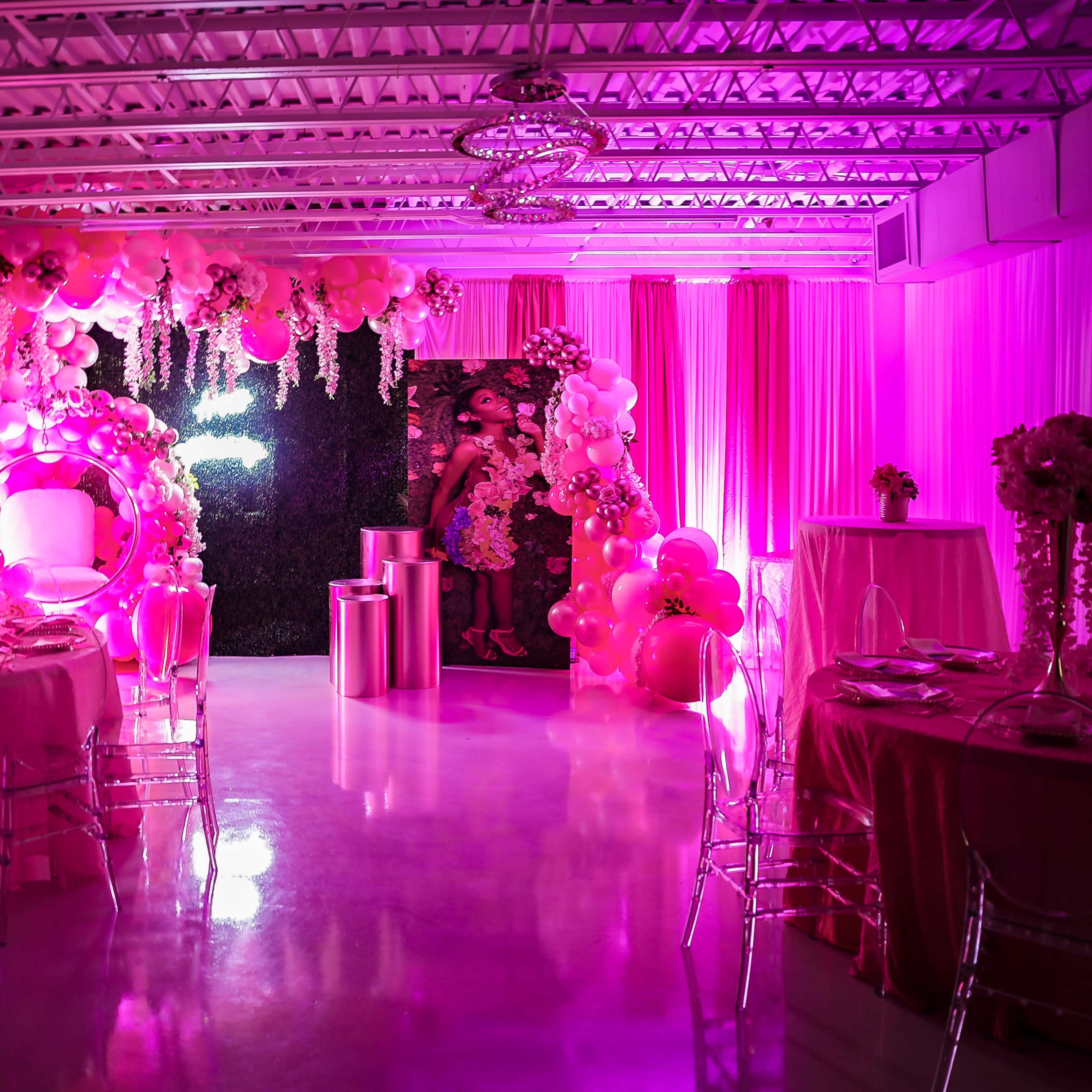 A decorated event space filled with pink balloons, floral centerpieces, and illuminated with soft pink lighting.