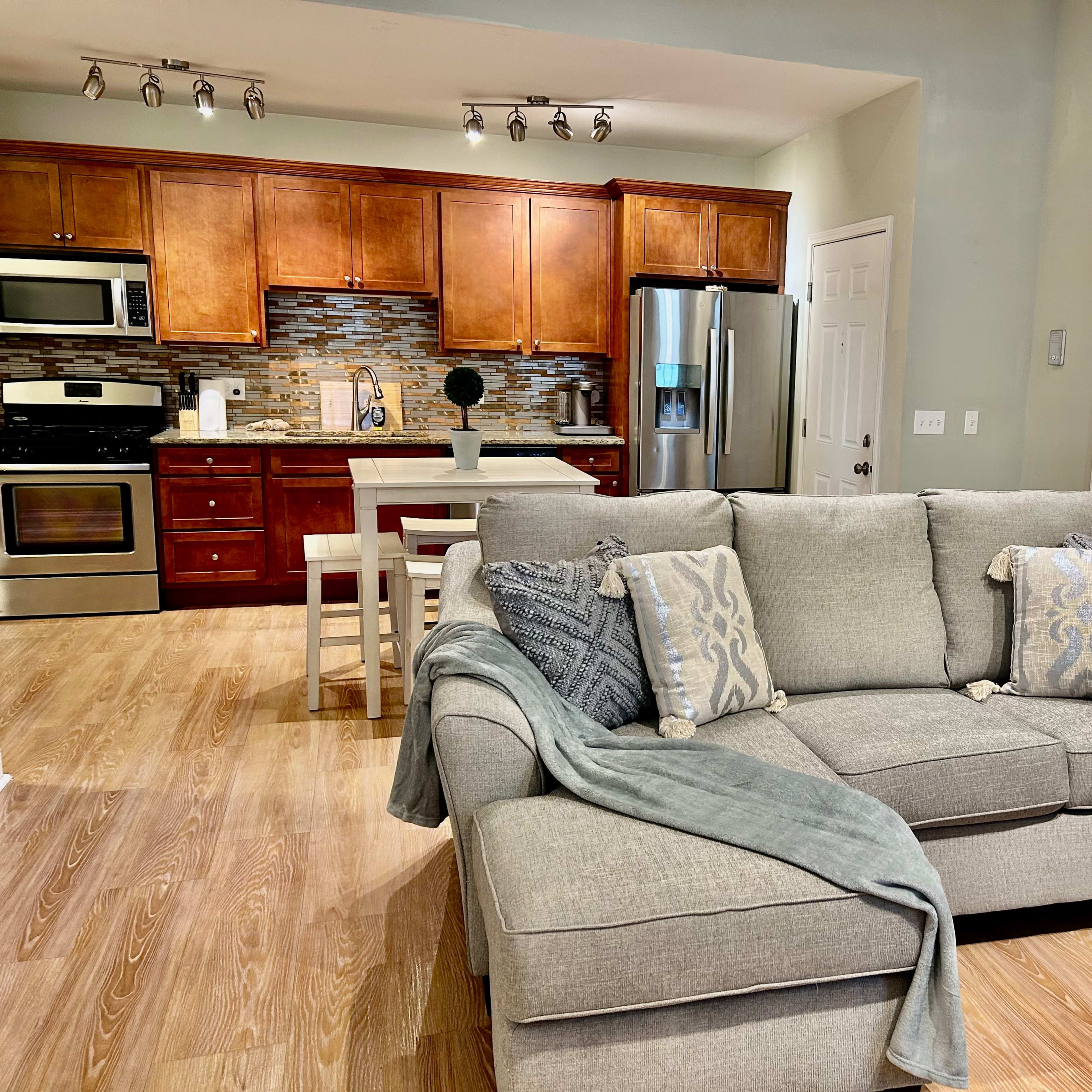 A living area with a gray couch adorned with decorative pillows, a wooden kitchen in the background featuring stainless steel appliances, and a dining table with chairs.