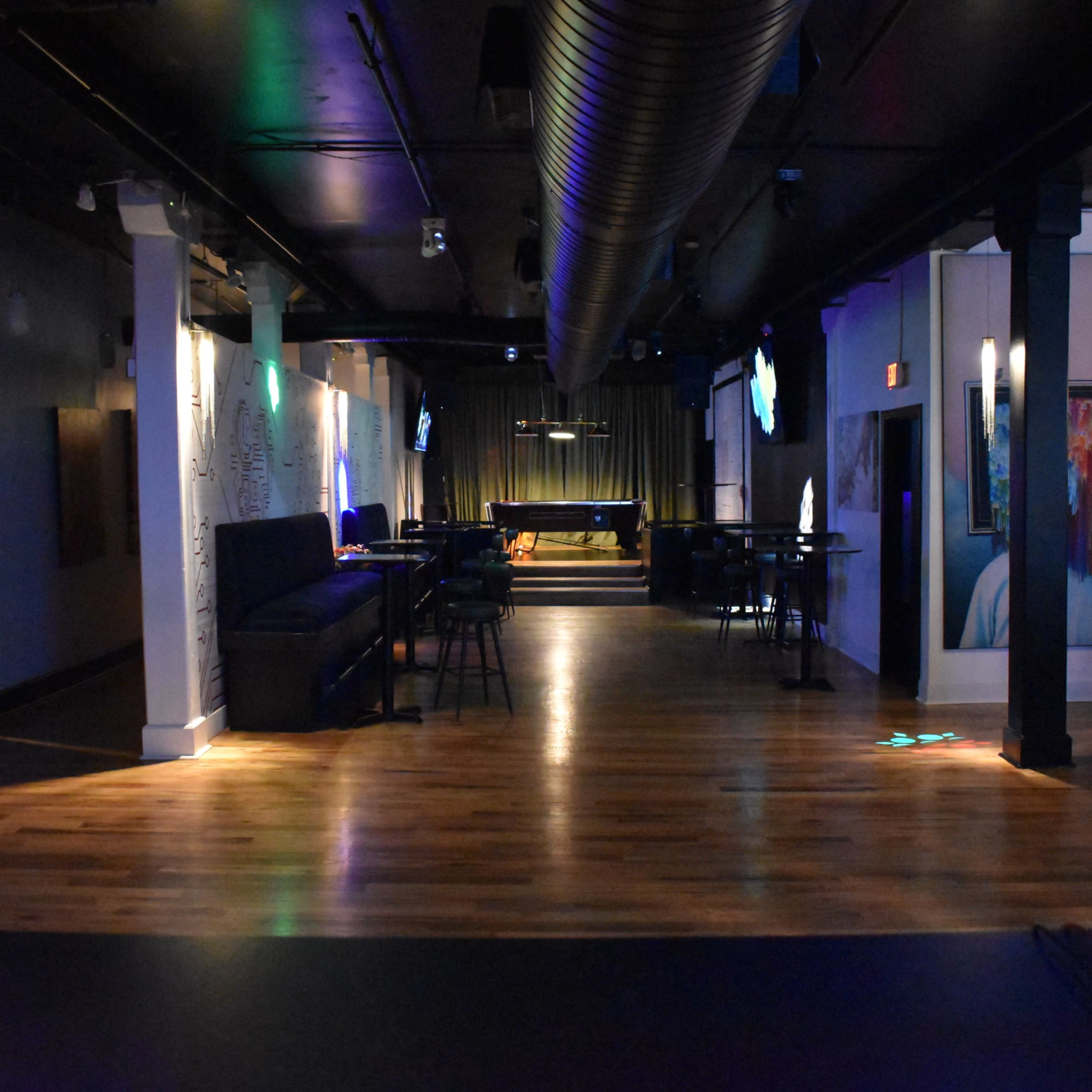 An empty bar or lounge area with wooden flooring, low seating along the walls, and a stage area at the far end.
