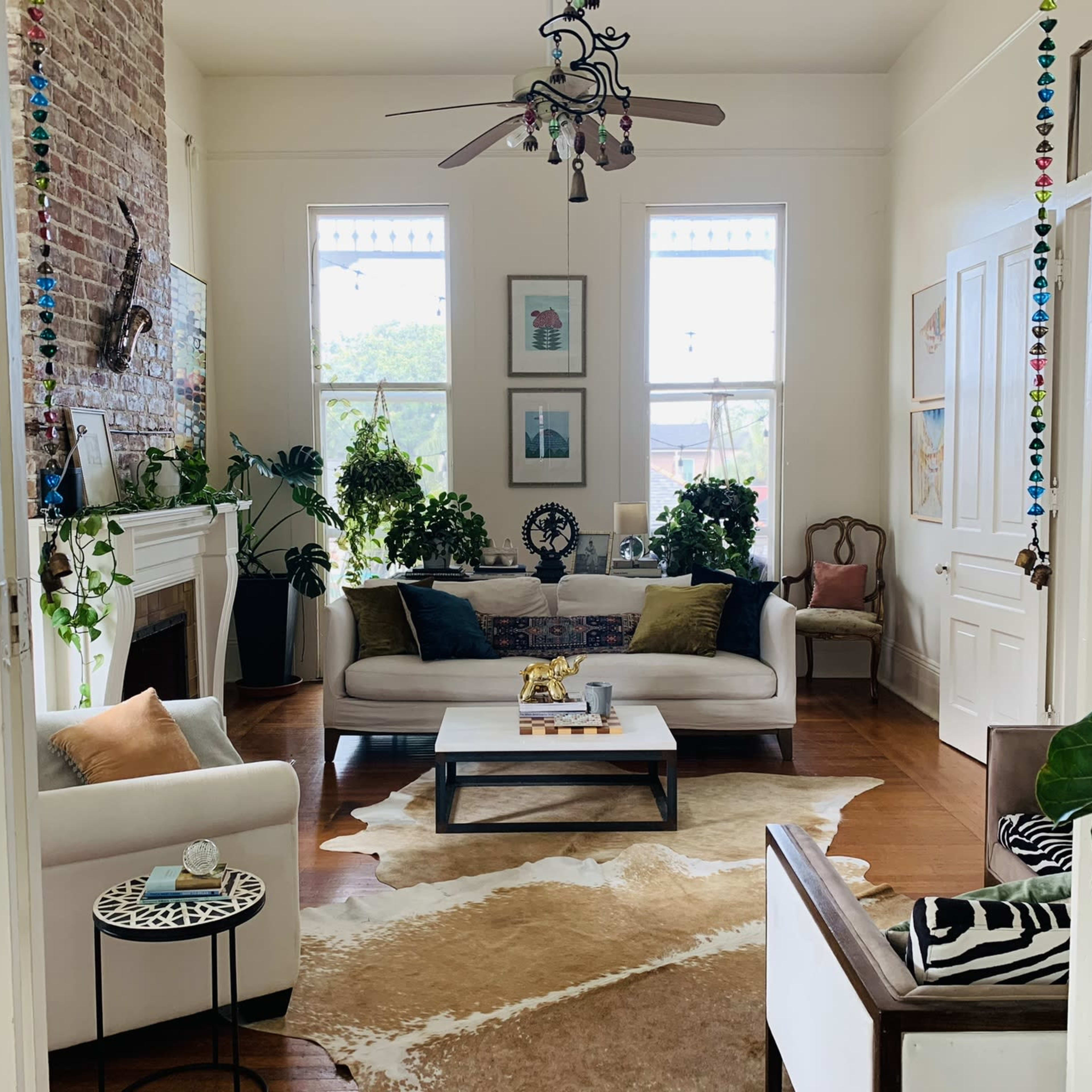 The living room features a combination of modern furniture, a large area rug, and several potted plants near bright windows.