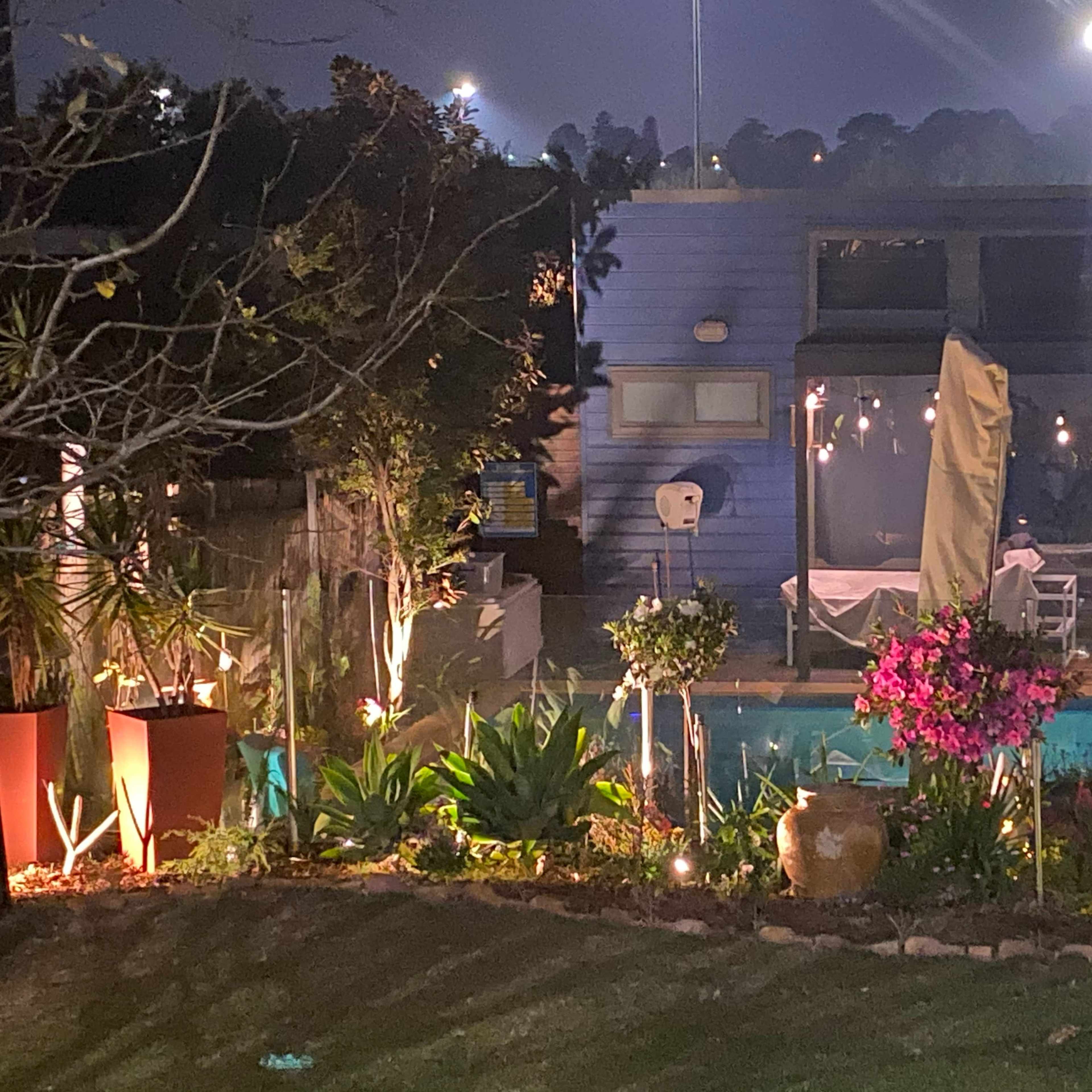 A backyard scene at night features a well-lit pool area surrounded by potted plants and a modern blue house.