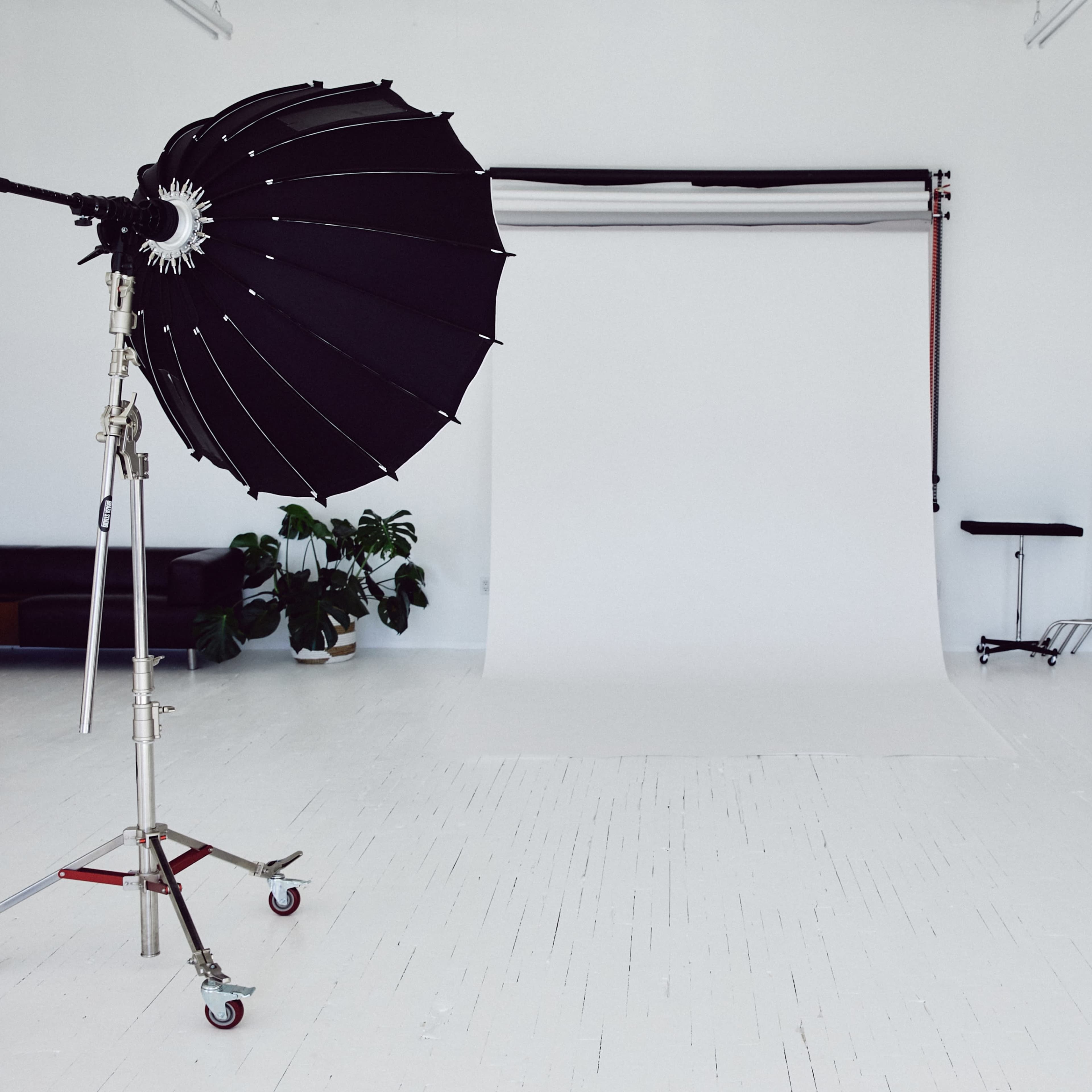 A photography studio features a large black umbrella light, a white backdrop, and various equipment on a clean, white floor.