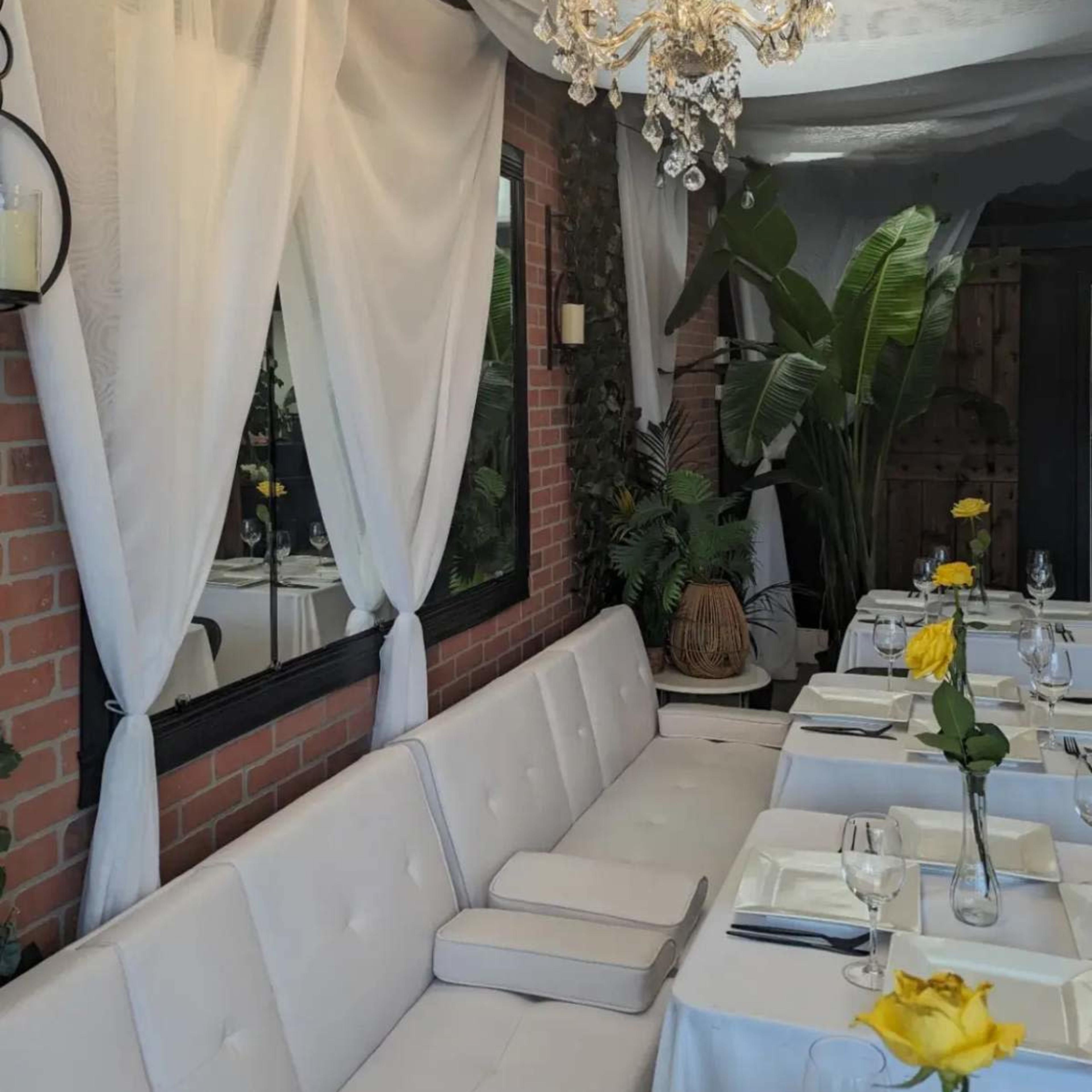 A restaurant interior featuring a white upholstered bench, tables set with plates and glasses, and plants placed throughout the space.