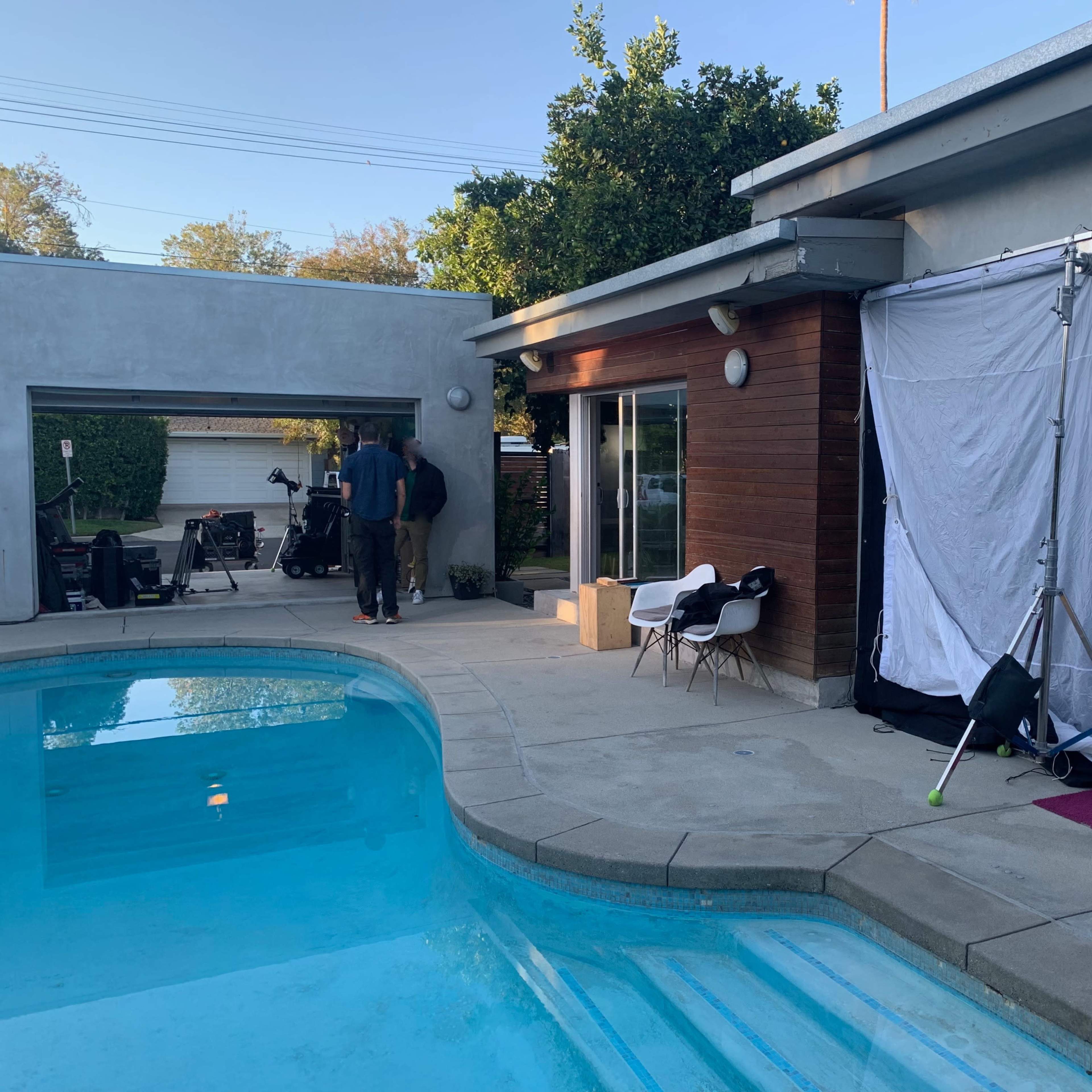 A film crew sets up near a swimming pool outside a modern house, with a large white backdrop partially propped up.