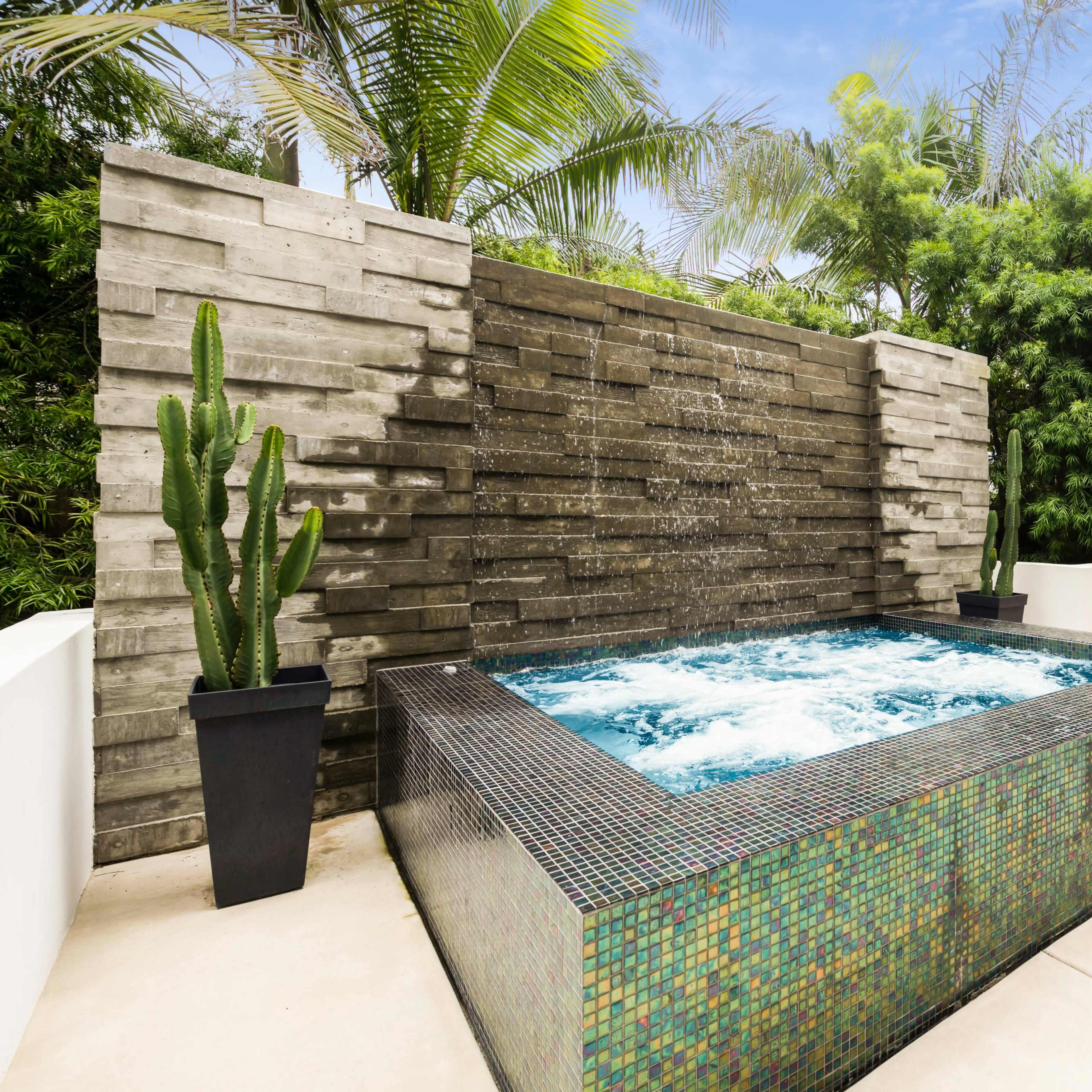A modern hot tub is situated next to a textured concrete wall with a water feature, surrounded by lush greenery and a dining area.