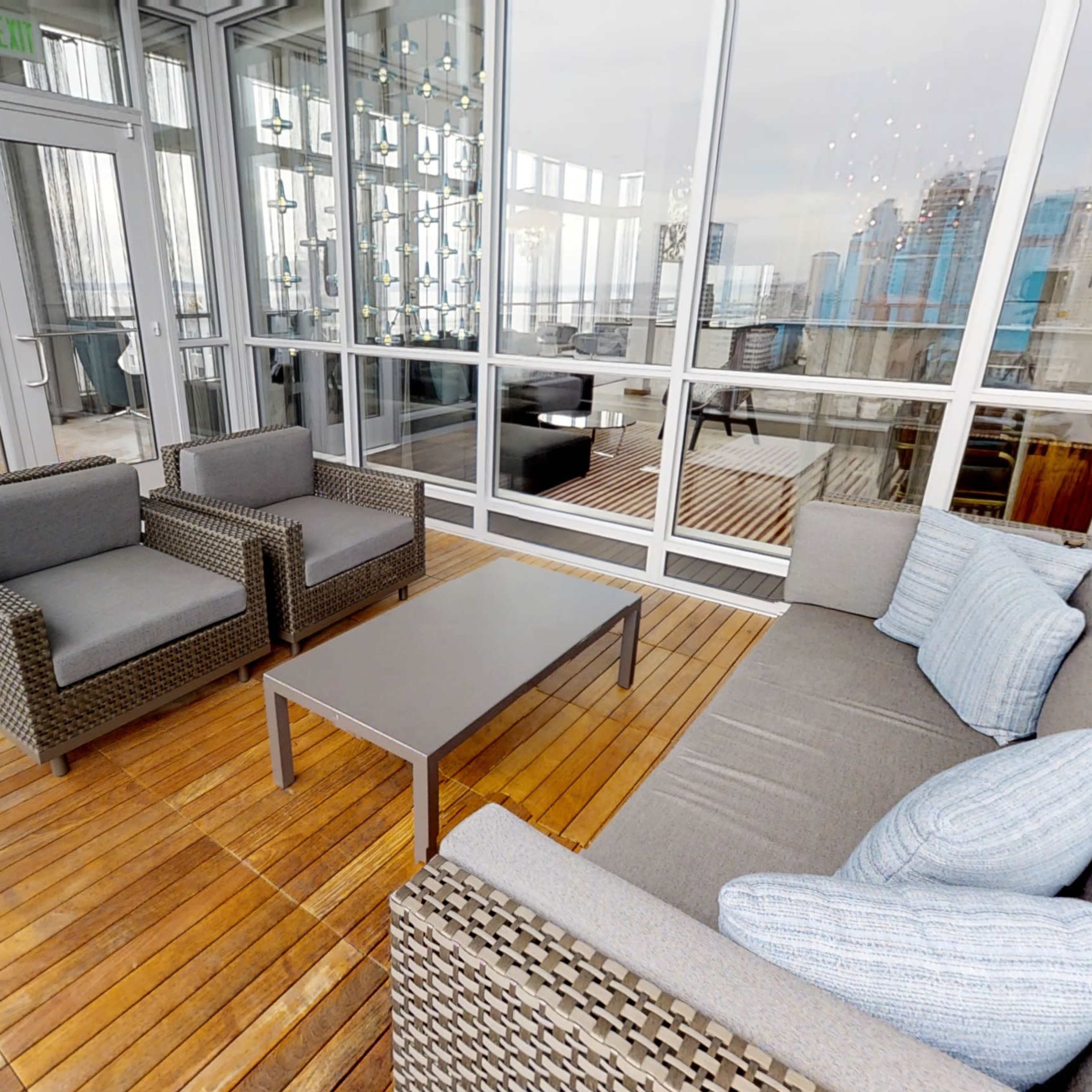 The scene shows a modern, furnished patio with two armchairs and a couch set around a coffee table, enclosed by glass walls offering a view of the city.