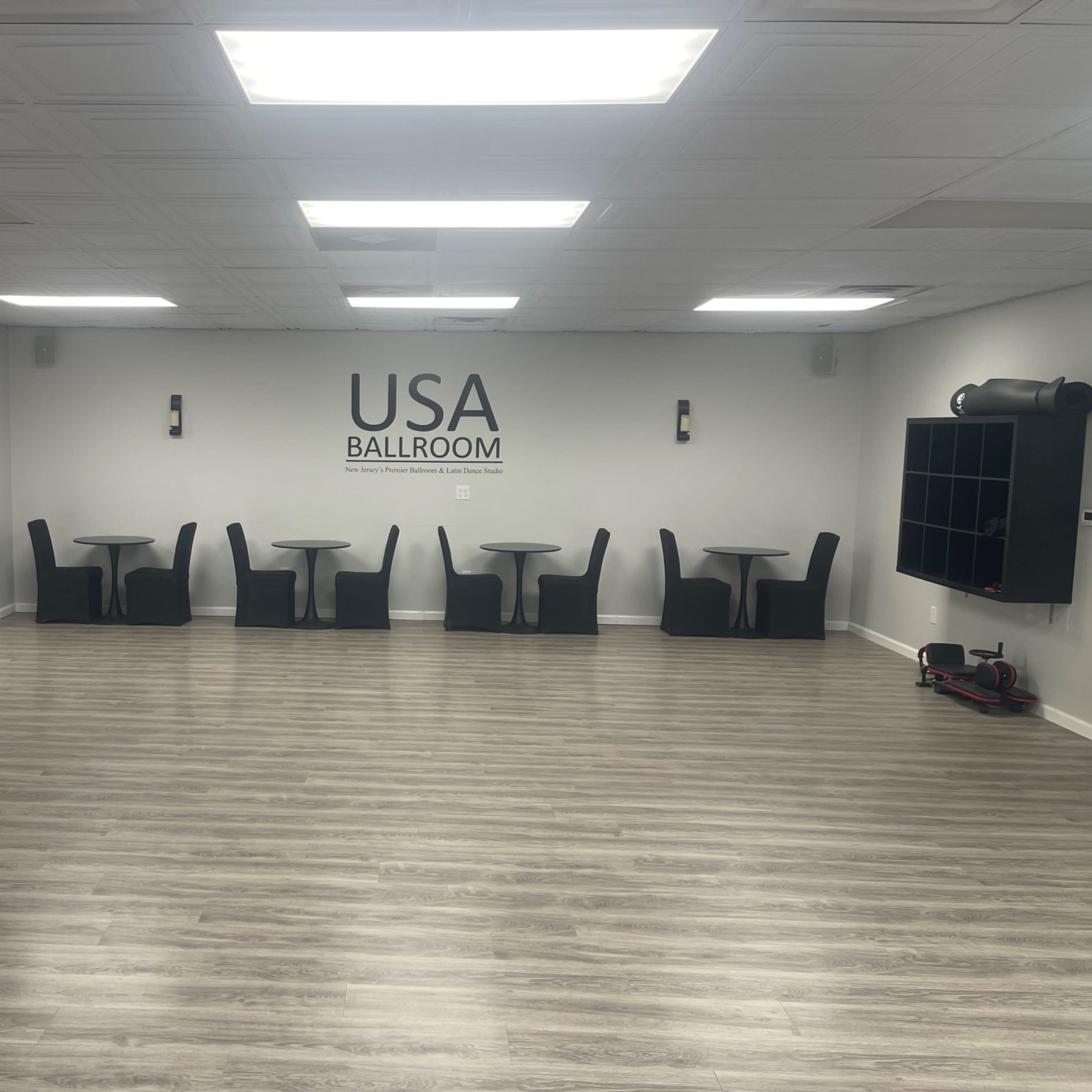 A spacious and empty ballroom with several round tables and chairs arranged around the room, featuring a large "USA BALLROOM" sign on the wall.