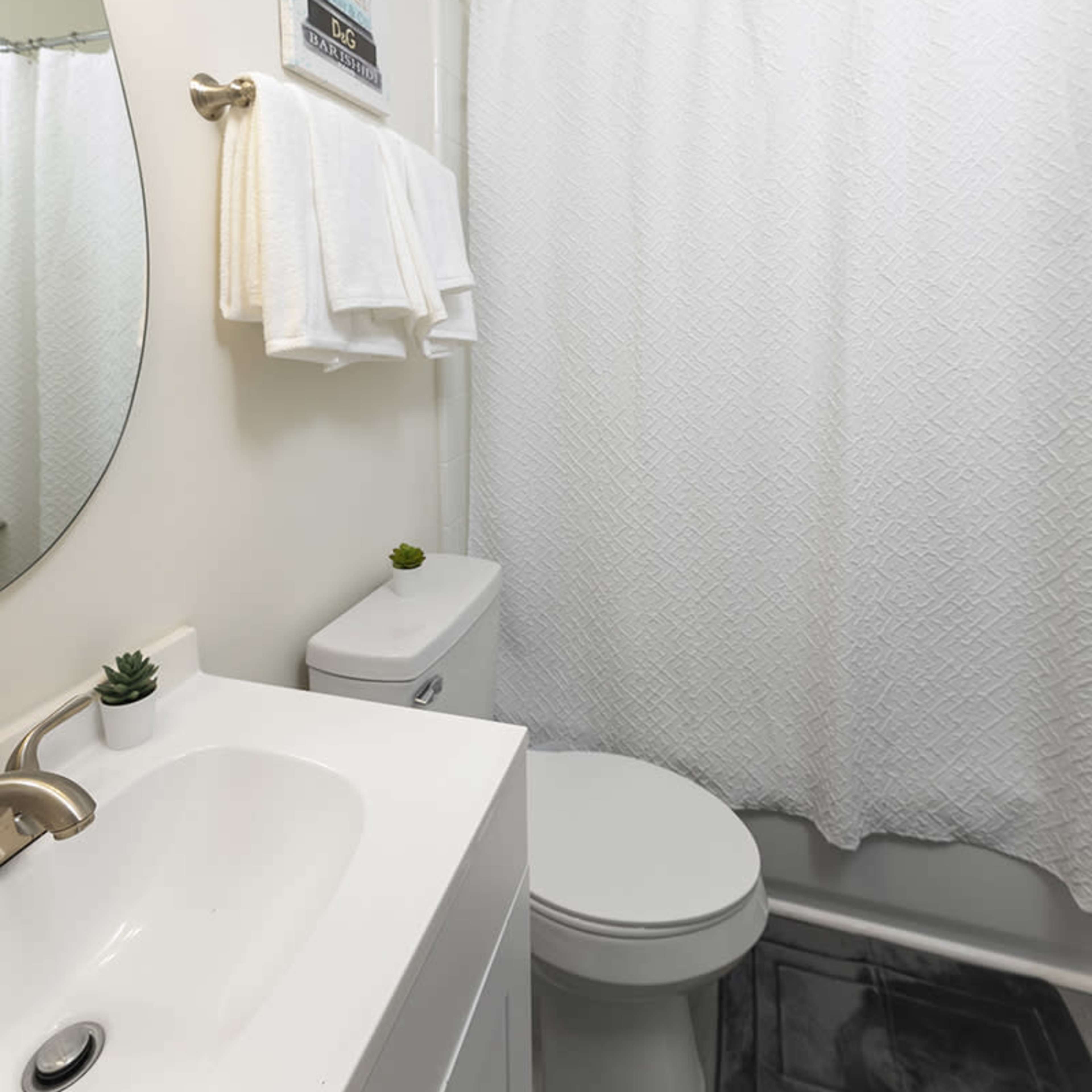 A clean bathroom featuring a white shower curtain, a circular mirror, a sink with a faucet, and neatly arranged towels.