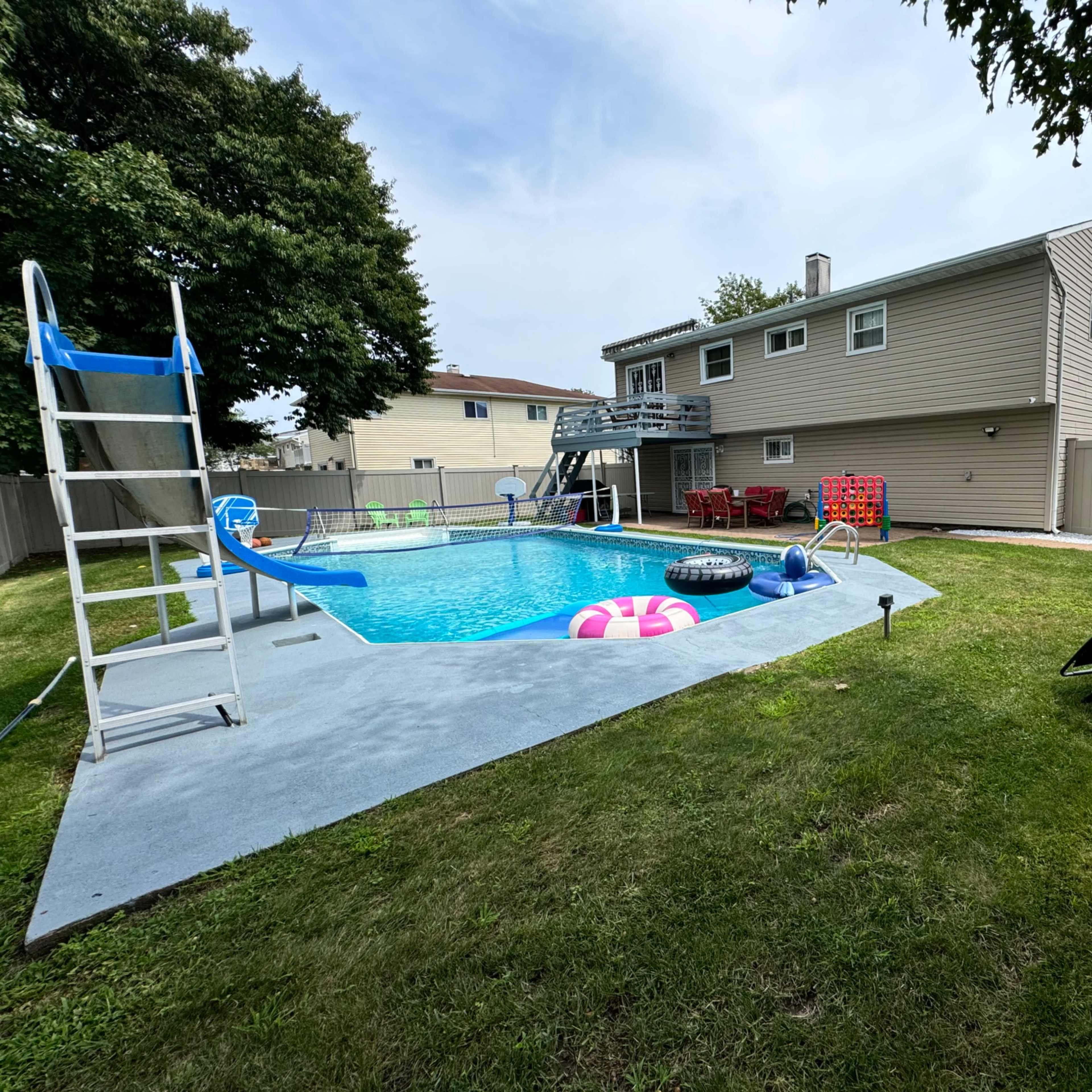 A backyard with a swimming pool, lounge chairs, and a small slide, surrounded by a fence and a two-story house in the background.
