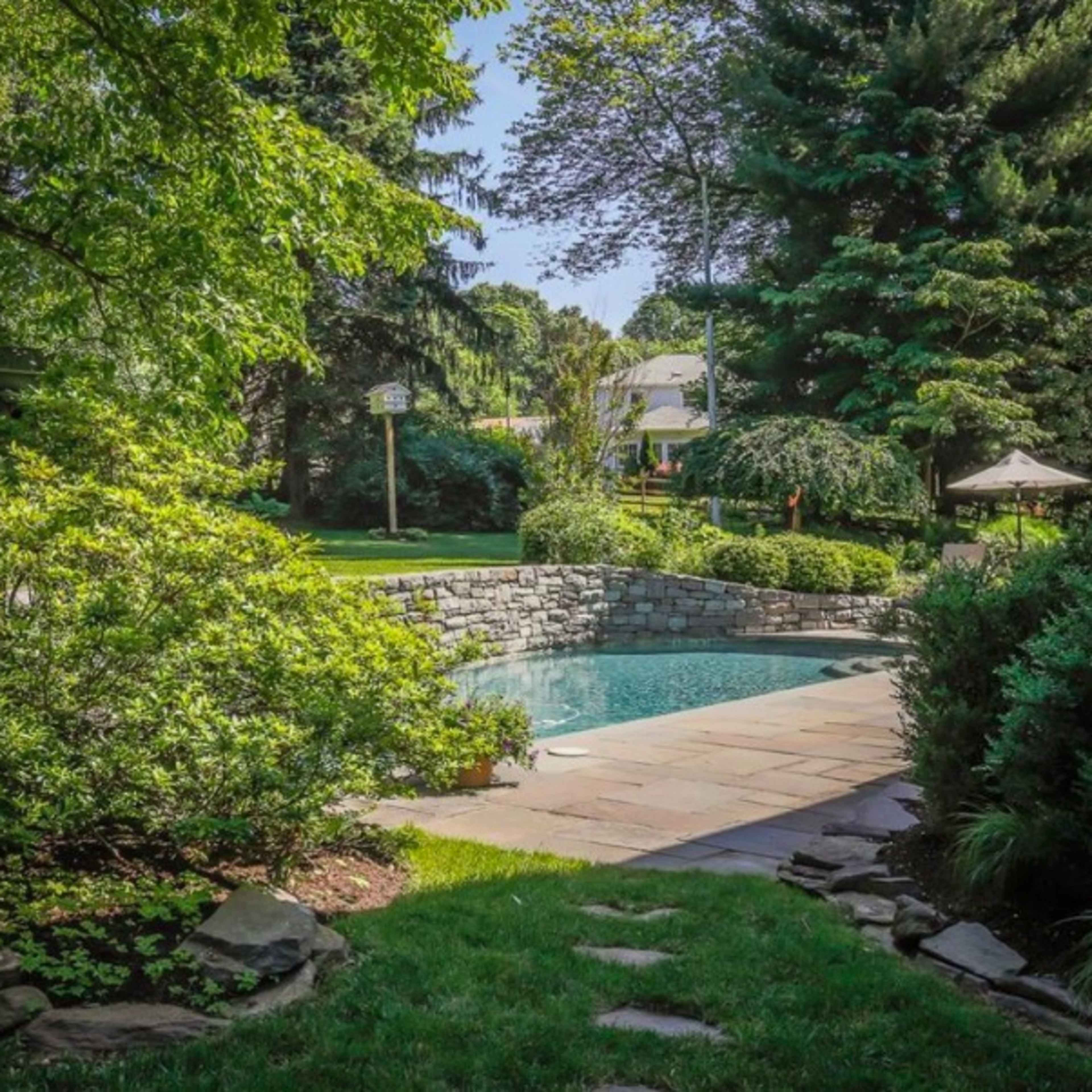 A landscaped backyard featuring a rectangular pool surrounded by stone patio, greenery, and trees.