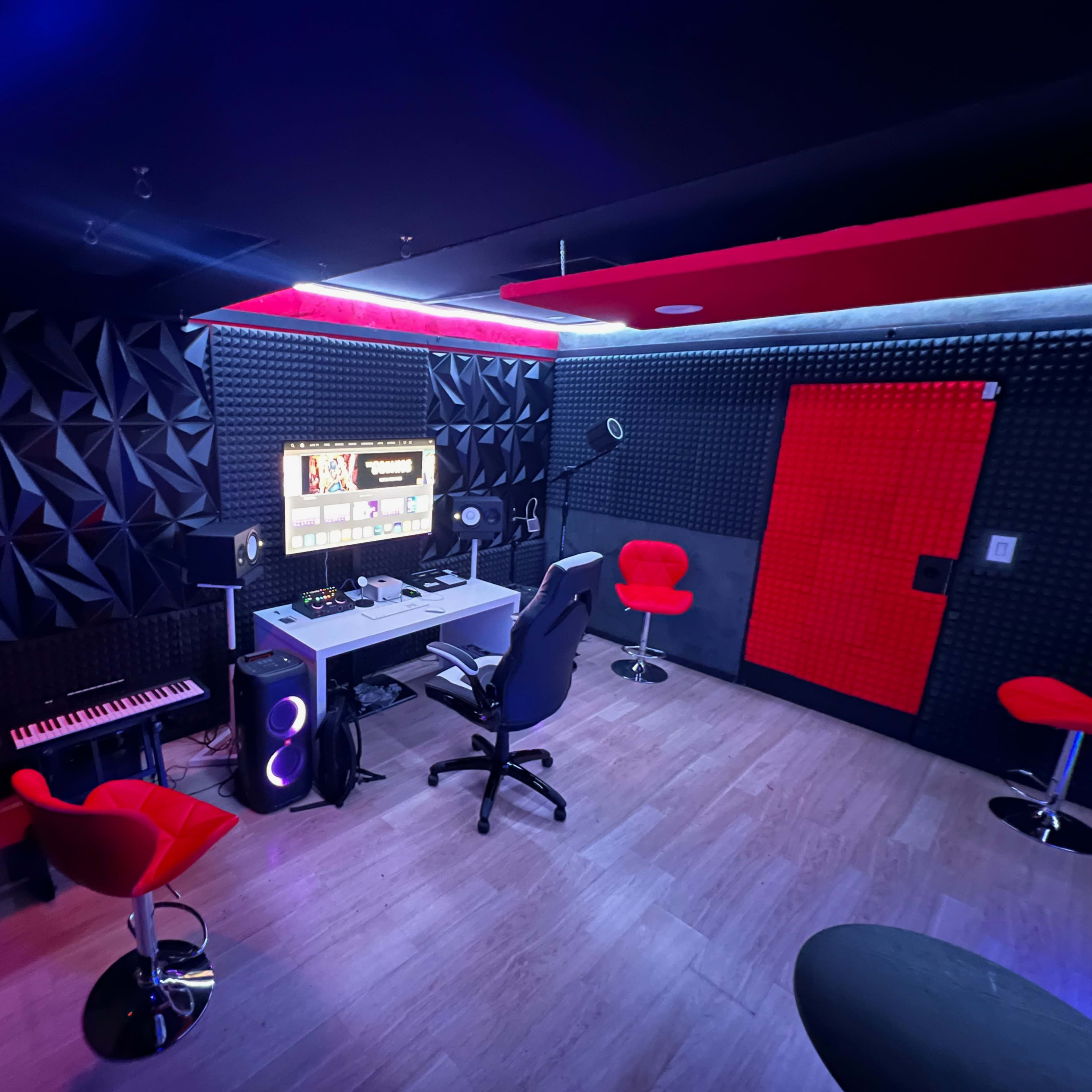 A modern recording studio featuring a central desk with a computer setup, red accent chairs, a keyboard, and soundproof walls.