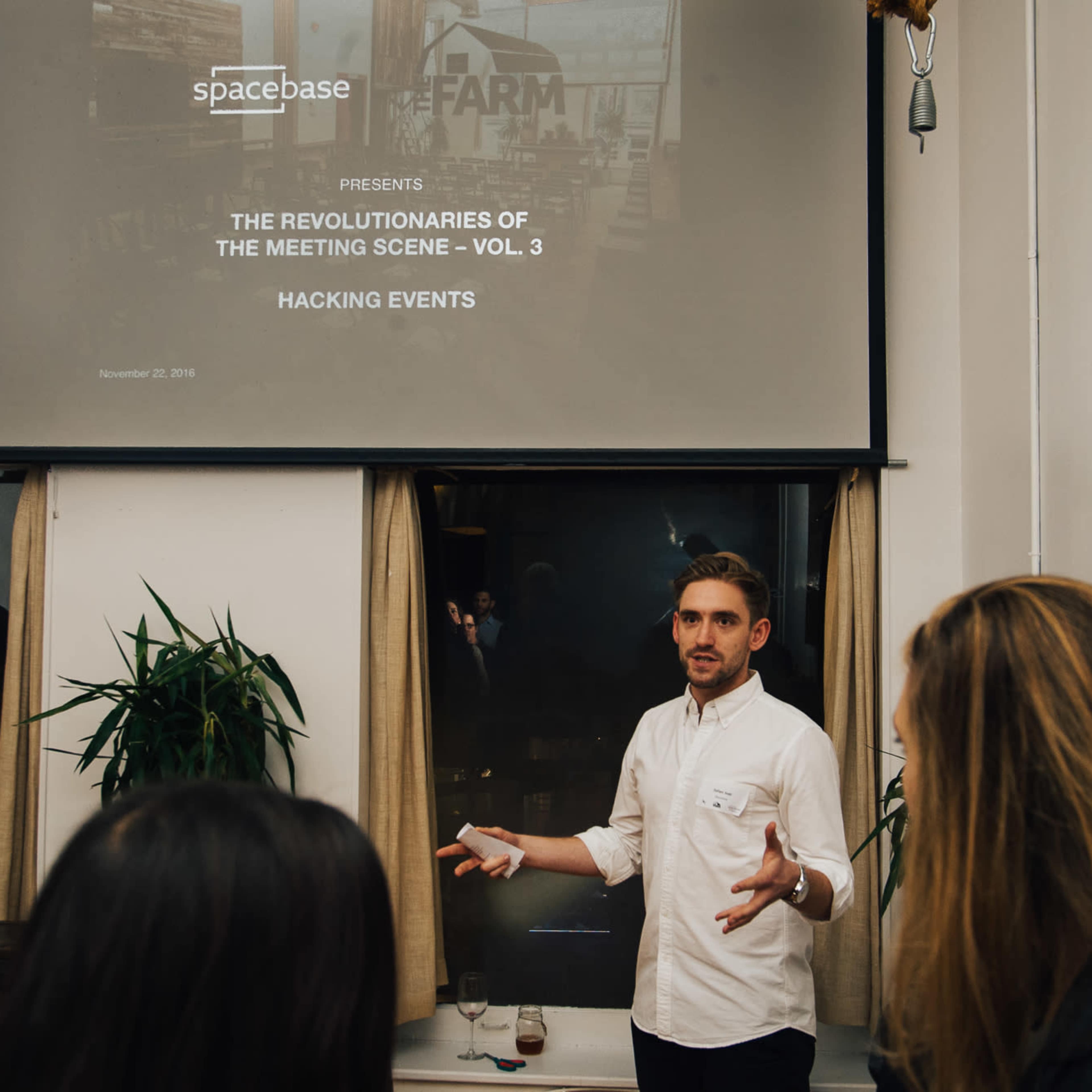 A speaker presents in front of an audience during a meeting event at a venue with a large screen displaying the event title.