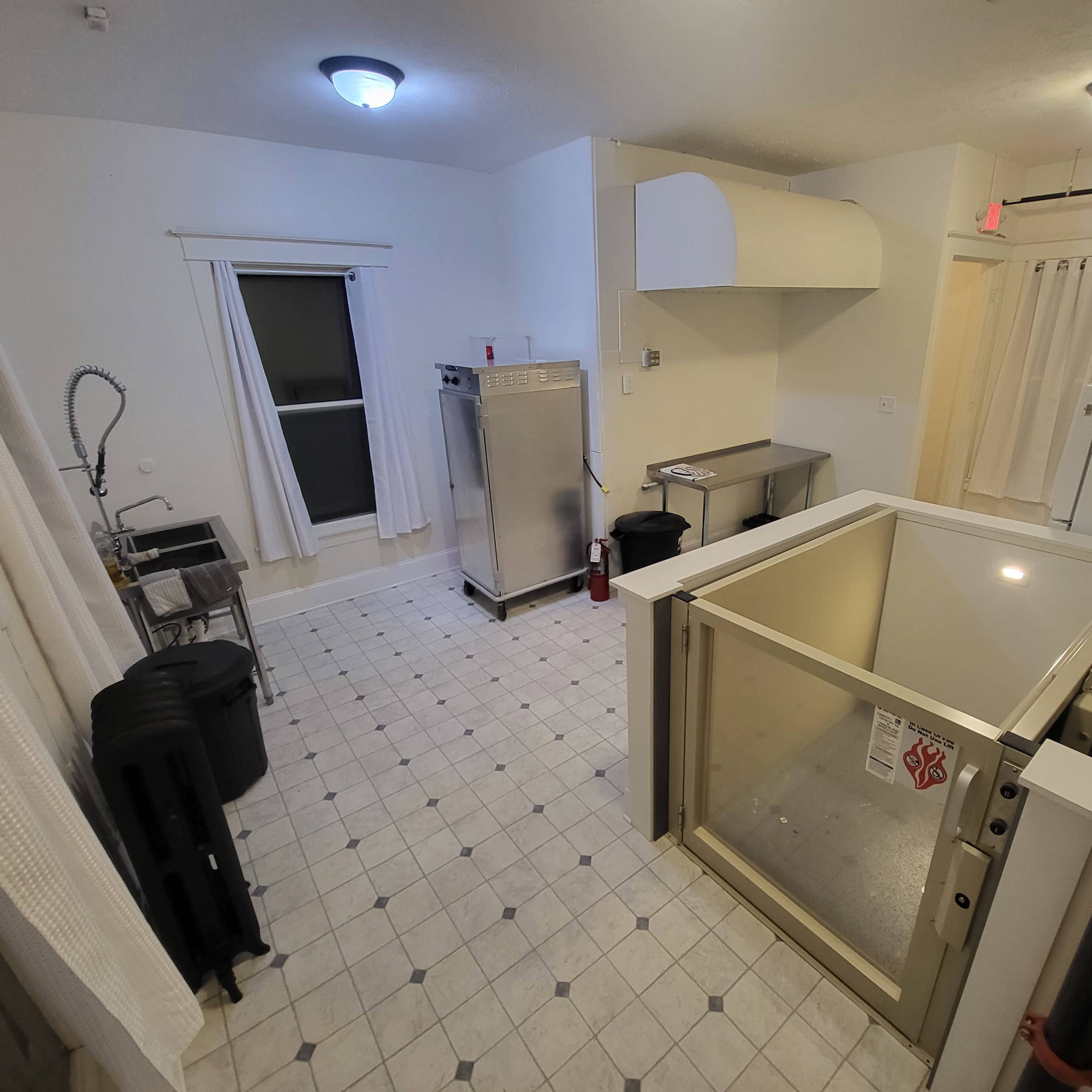A kitchen space with a stainless steel counter, a large freezer unit, and a window, all featuring tiled floors and white walls.