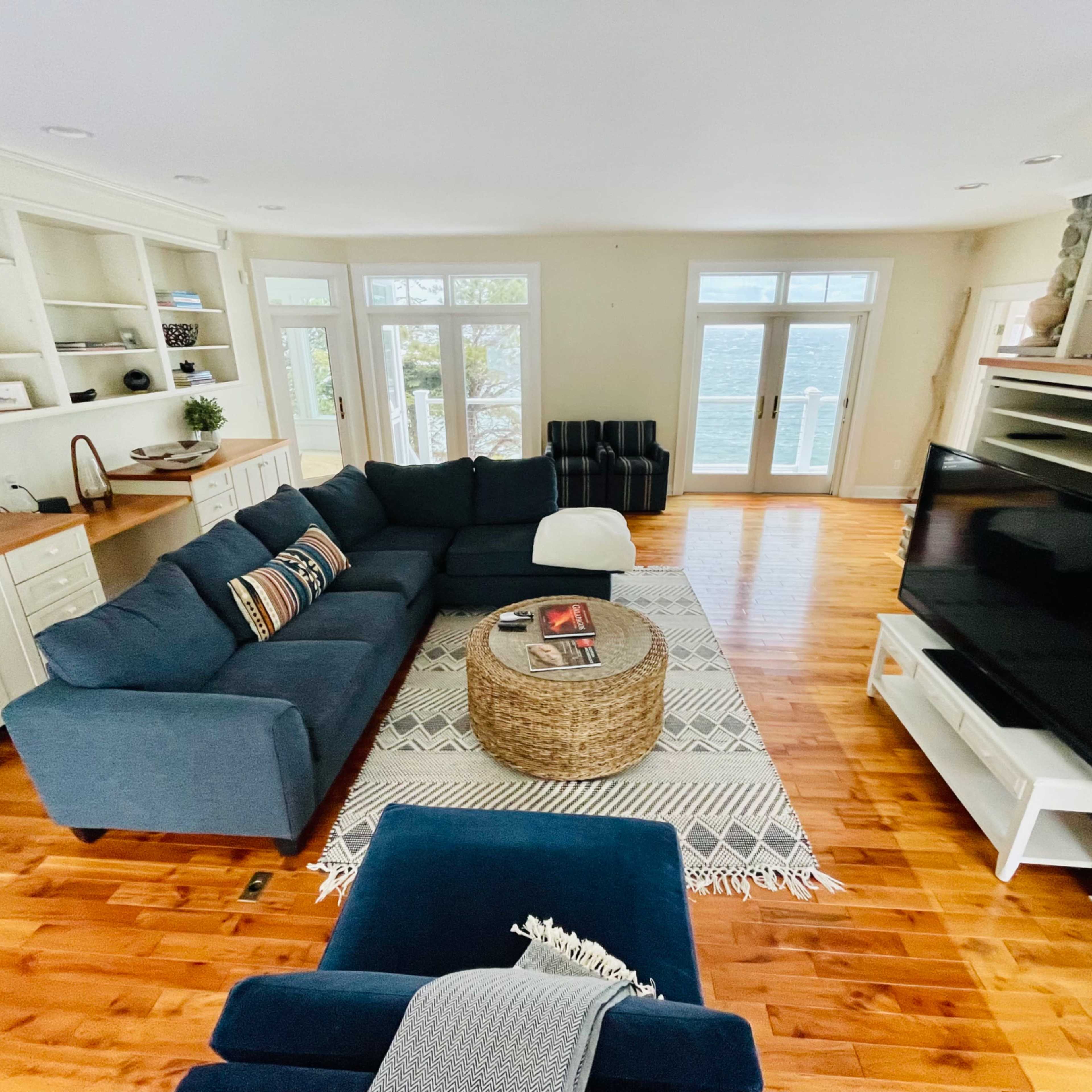 A spacious living room with a large blue sectional sofa, a round coffee table, and a television against a cream-colored wall, illuminated by natural light from large windows.