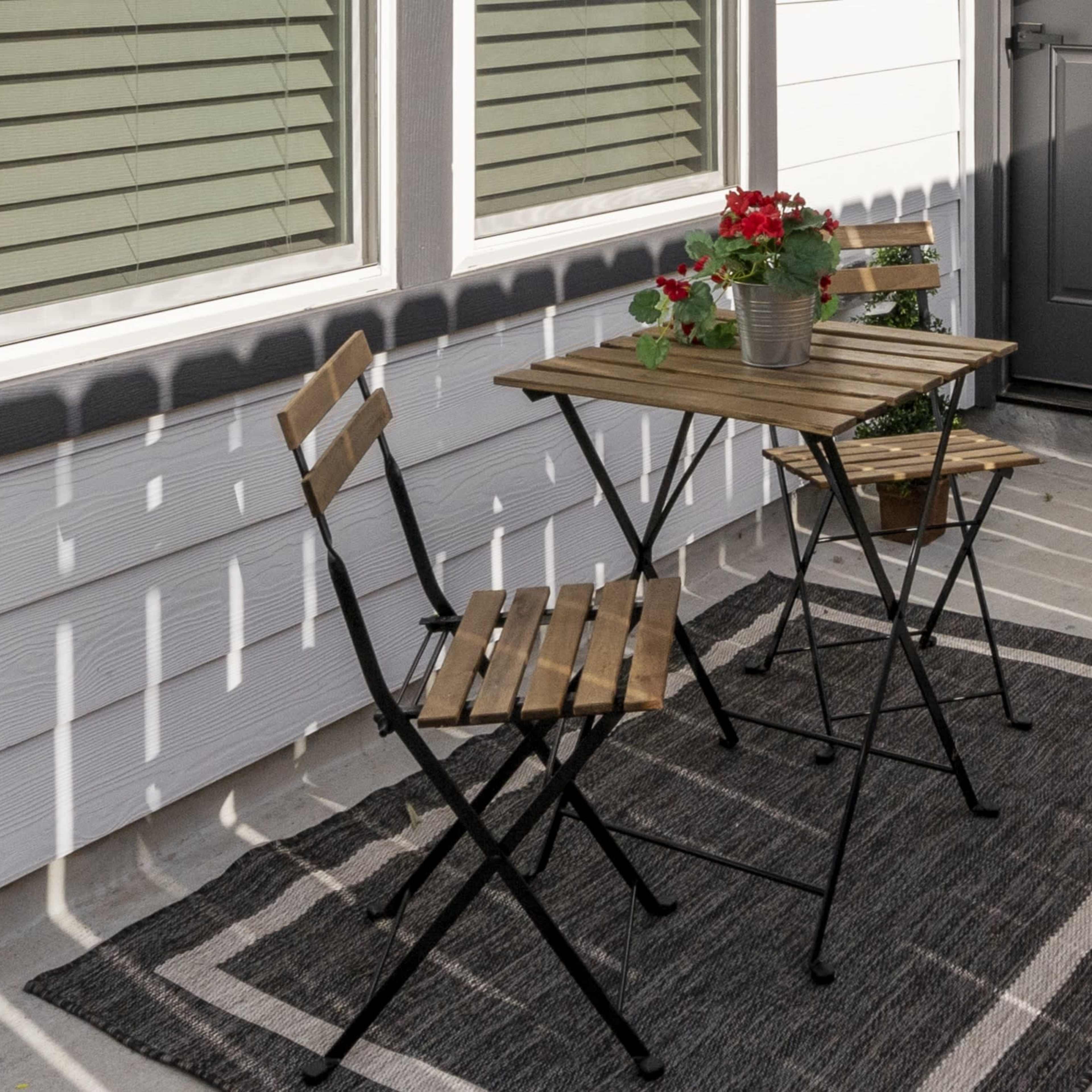 A small patio features a wooden table with two folding chairs and a potted plant surrounded by patterned outdoor rug.