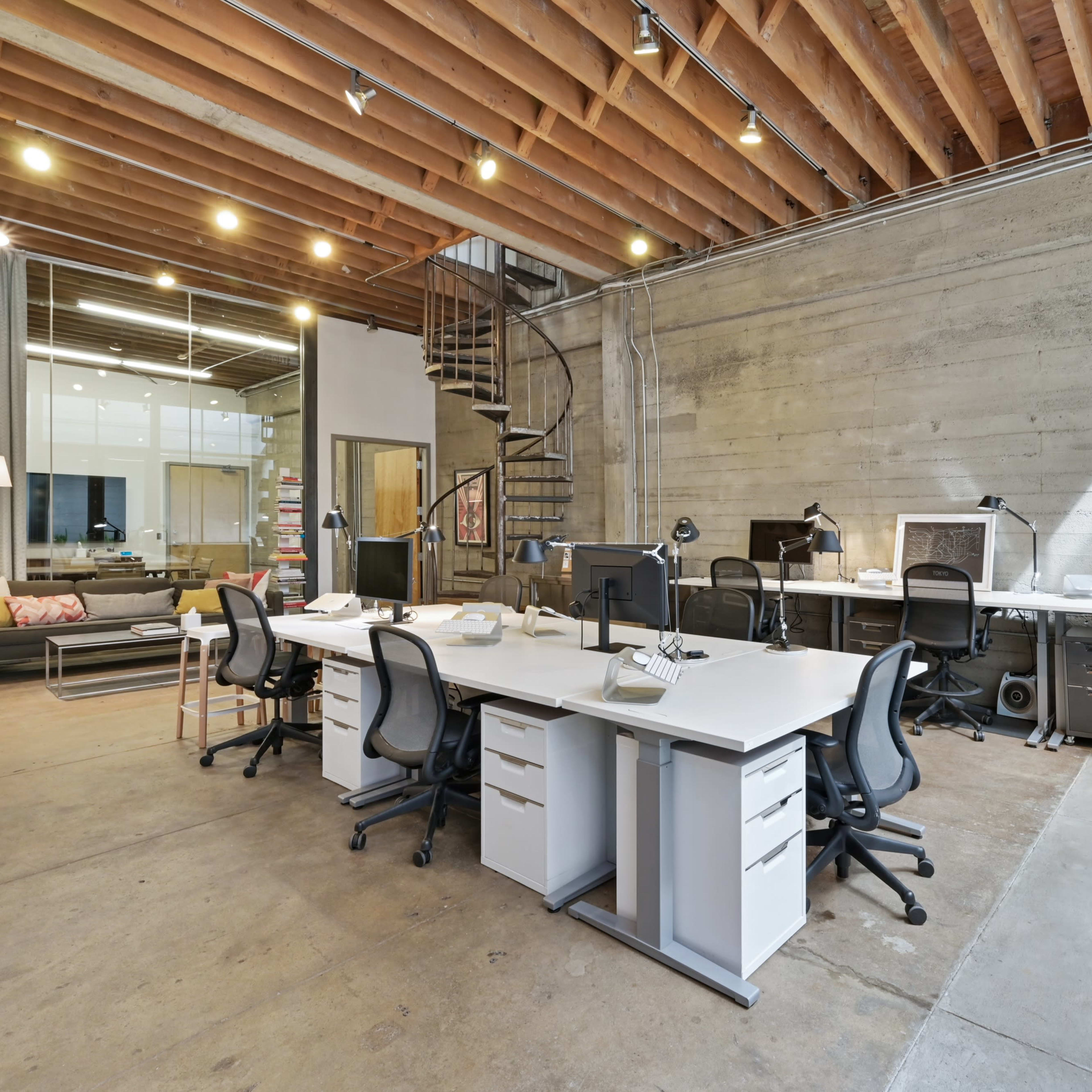 A modern office space with several white desks equipped with computers, a spiral staircase, and industrial-style exposed wooden beams and concrete walls.