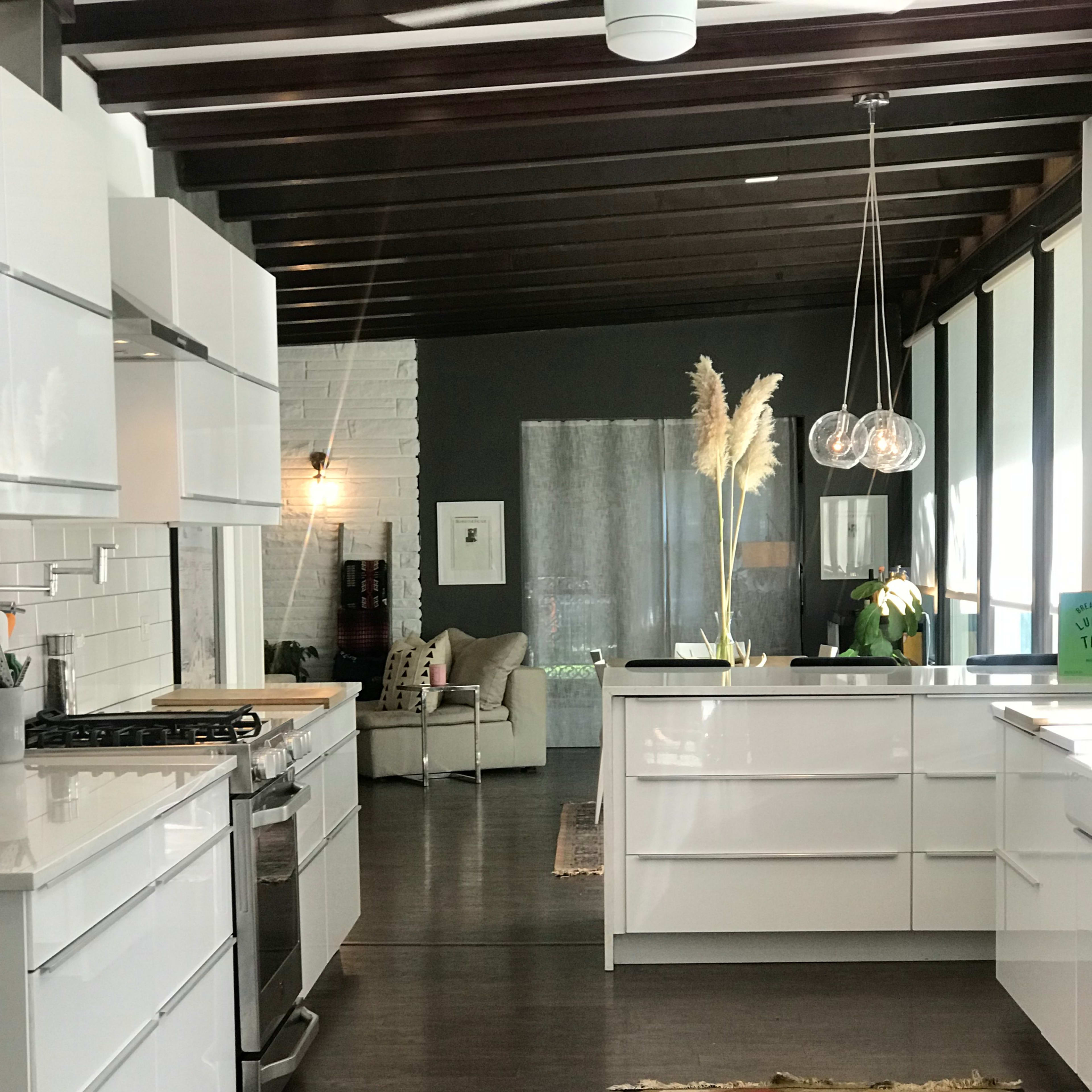 A modern kitchen features white cabinetry, a stainless steel stove, and hardwood floors, with a living area visible through large windows.