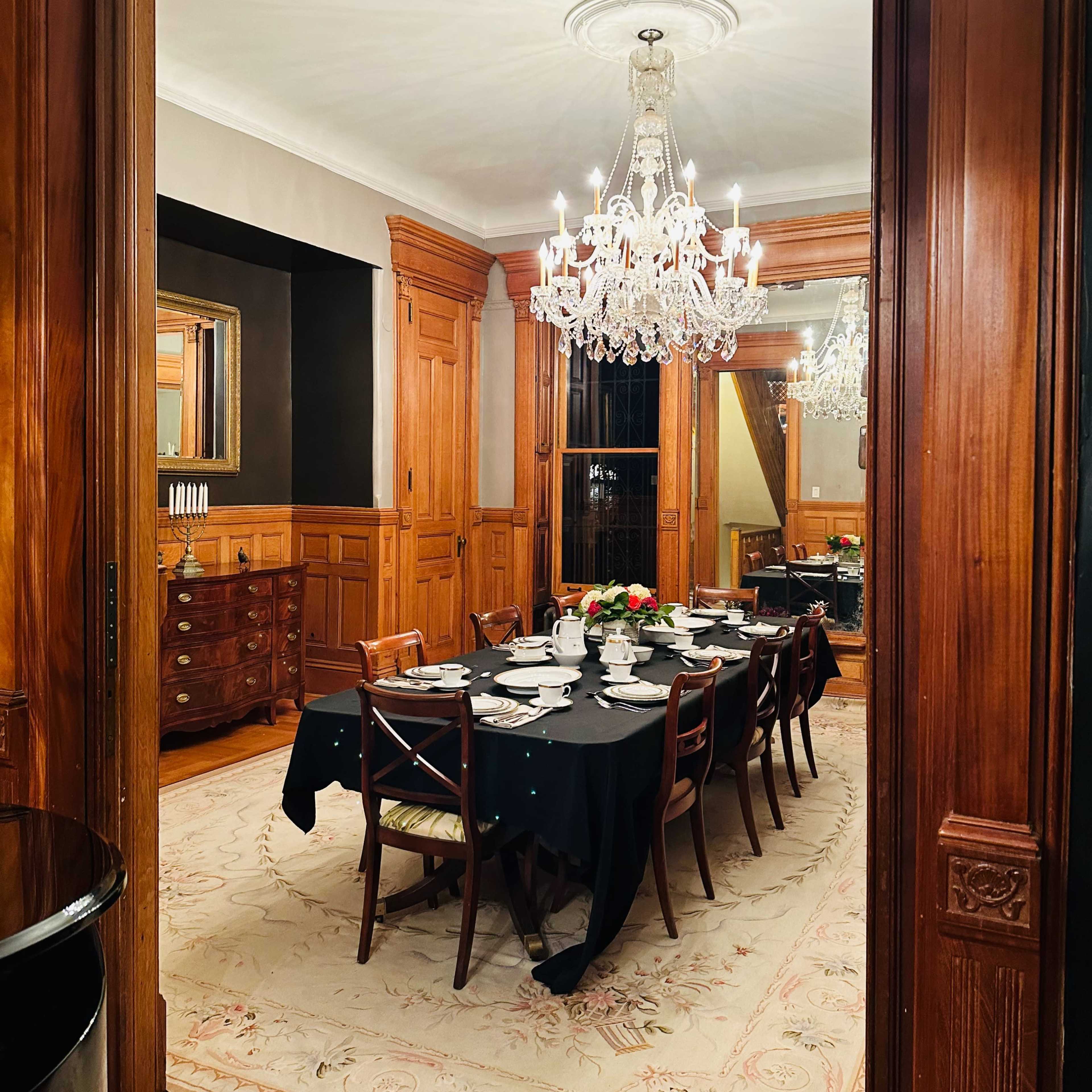 A formal dining room features a long table set for dinner, illuminated by a chandelier, and surrounded by wooden walls and furniture.