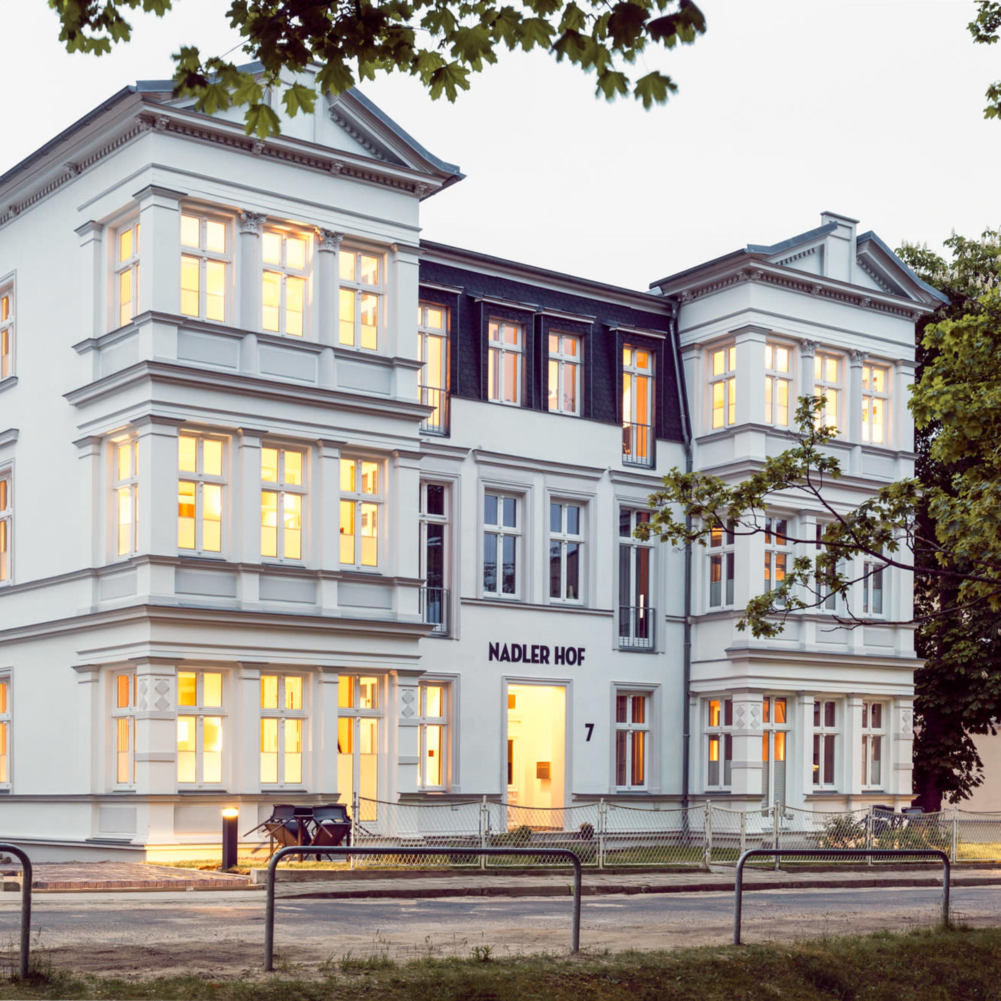 A large, white, multi-story building with numerous windows, illuminated from within, located at "Nadler Hof 7."