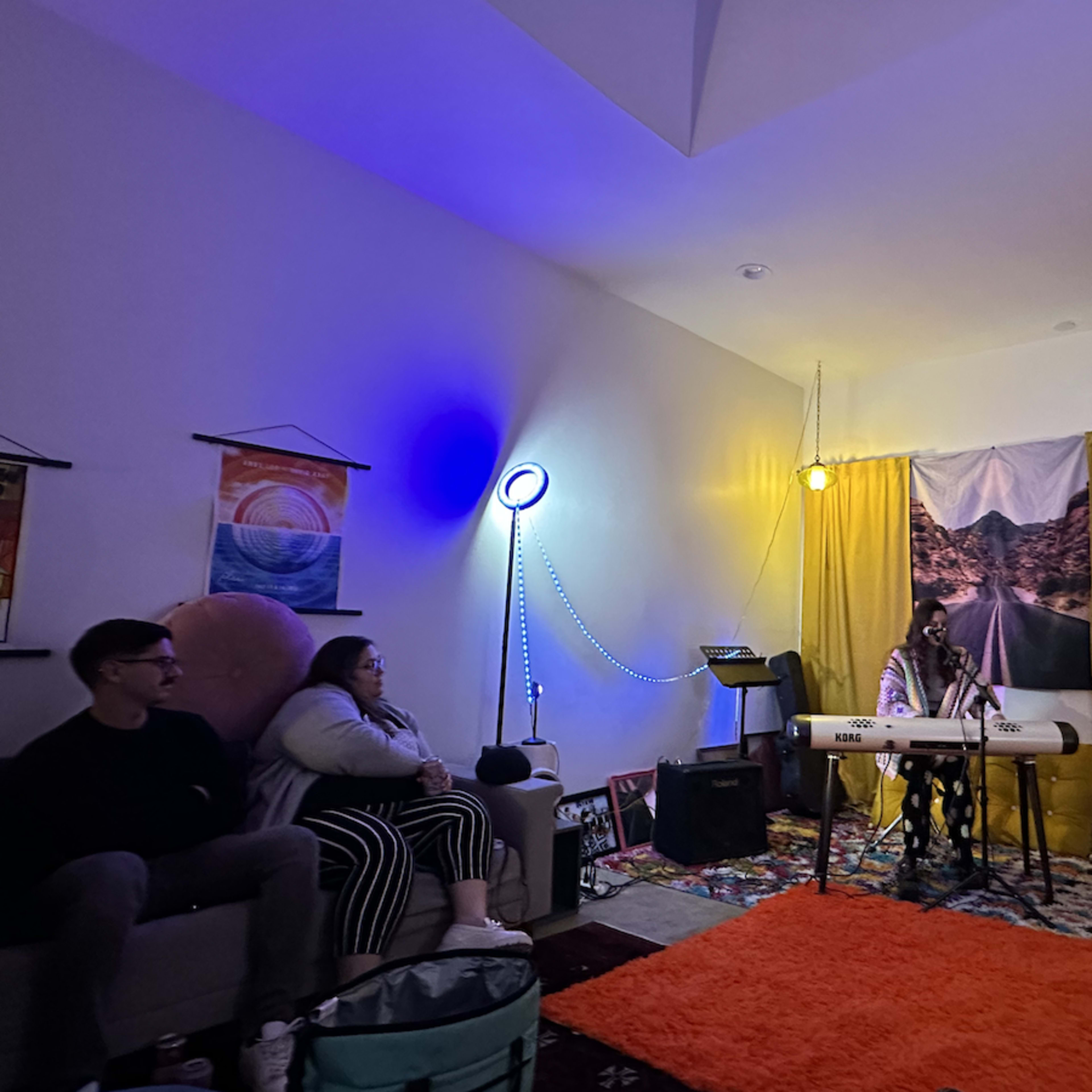 A musician performs at a small indoor gathering, with colorful lighting and a cozy seating arrangement.