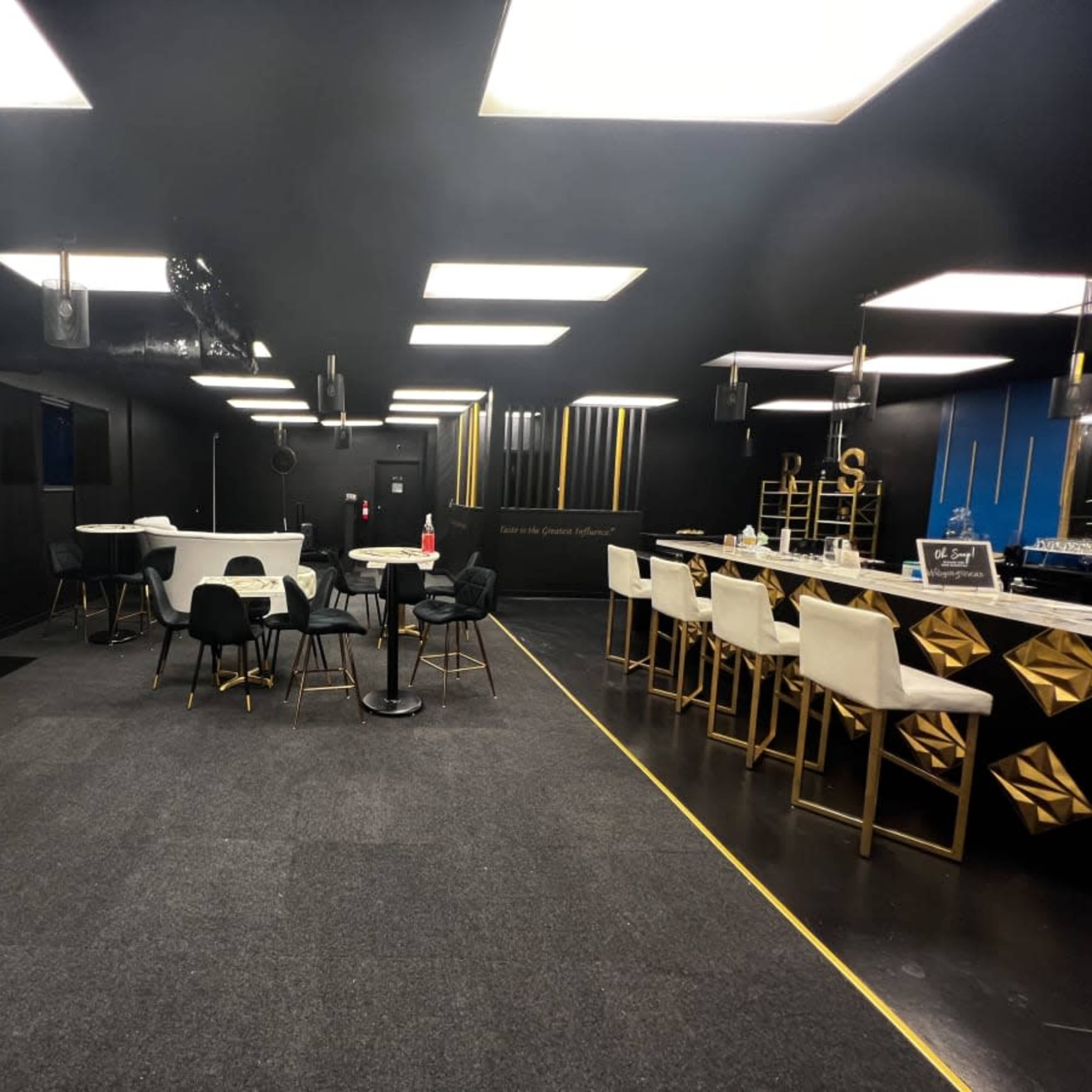 A modern, black-walled interior space featuring a mix of tables and chairs, a bar area, and decorative elements like plants and lighting fixtures.