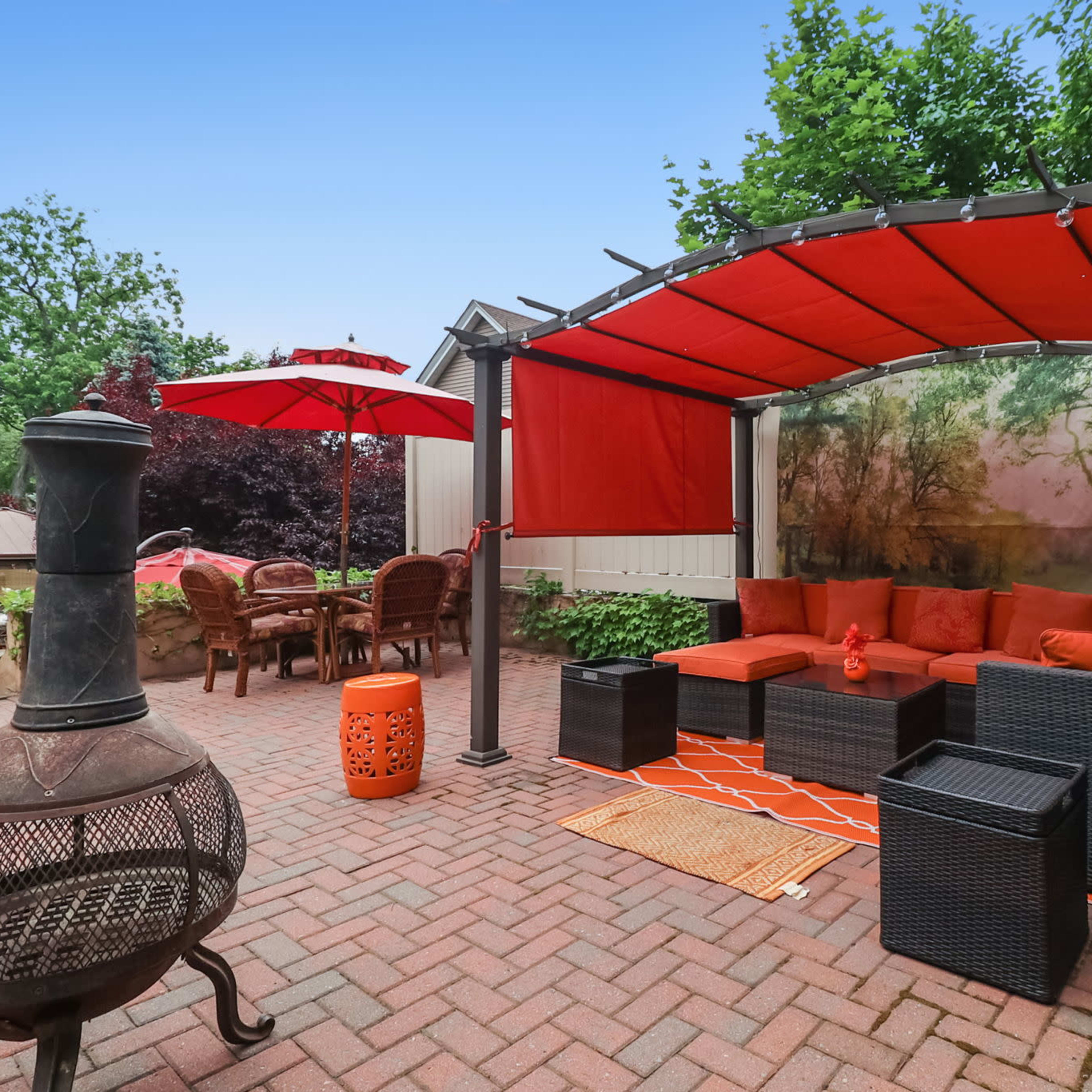 The patio features a covered seating area with orange cushions, a matching rug, a fire pit, and a dining set beneath an umbrella.