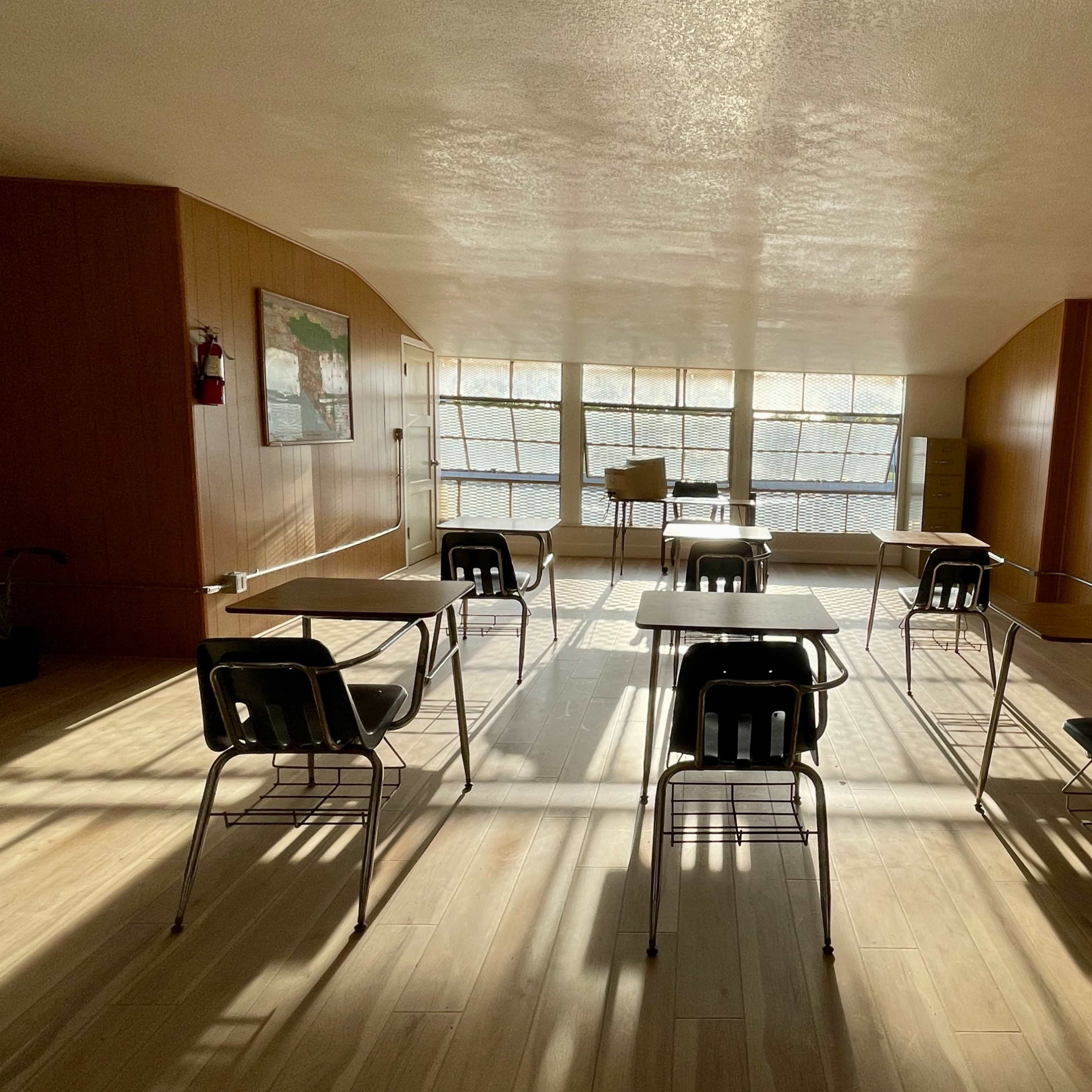 An empty classroom with rows of desks and chairs arranged under large windows, which allow natural light to illuminate the room.