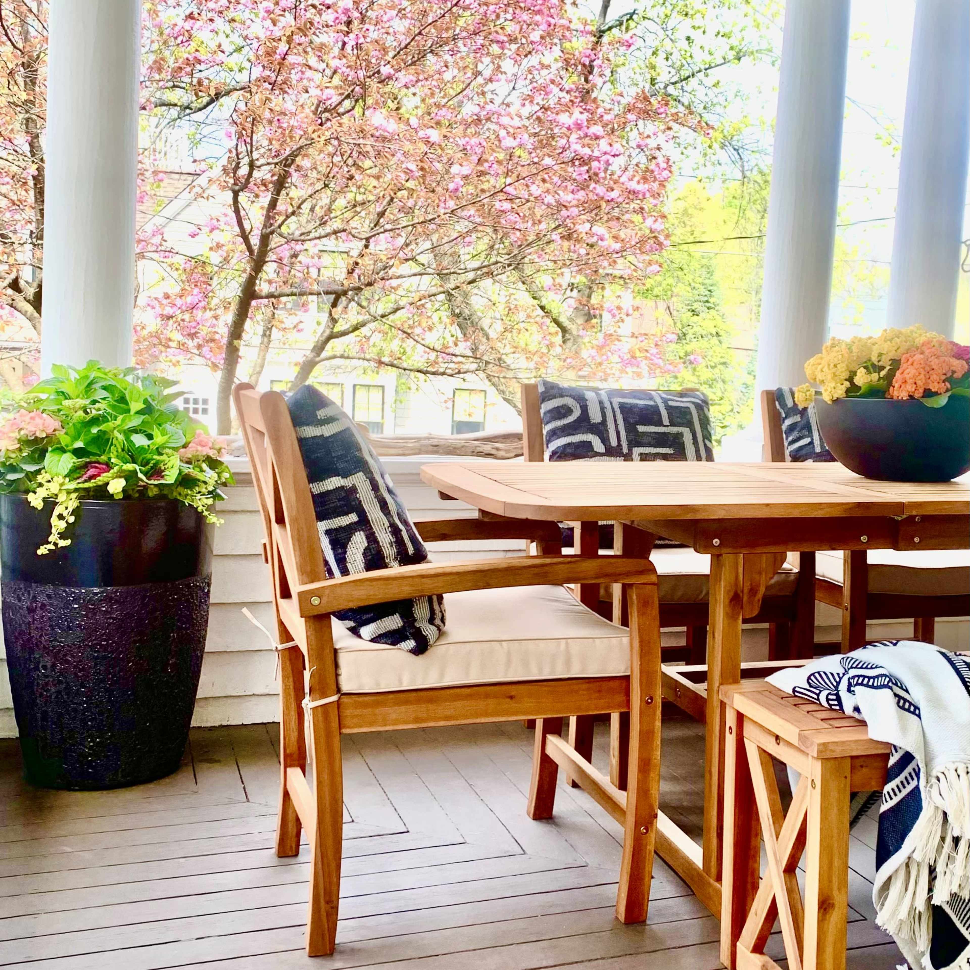 A wooden dining table with chairs is set on a porch, surrounded by potted plants and blooming trees.