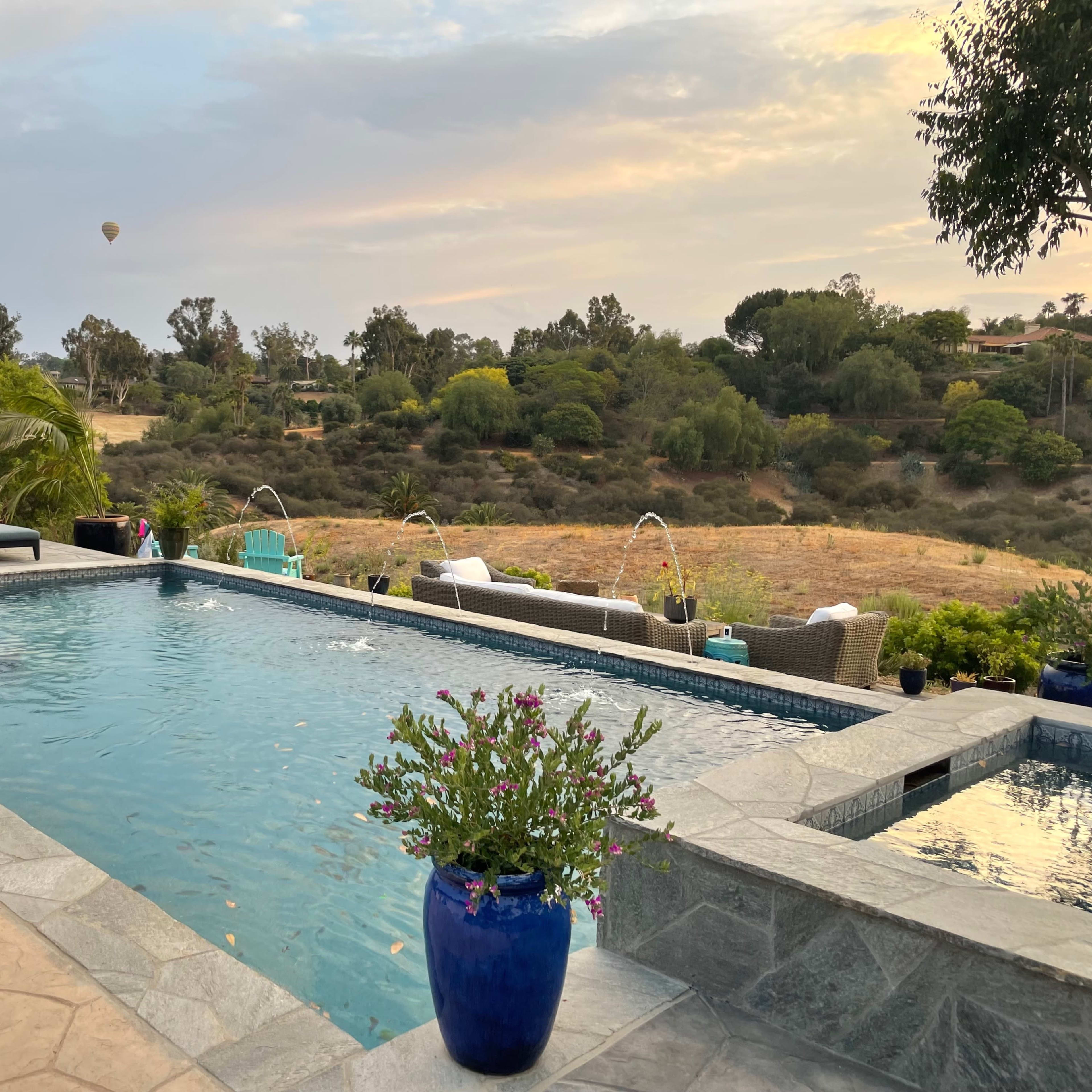 A pool area with lounge chairs, surrounded by greenery and a distant hot air balloon in the sky.