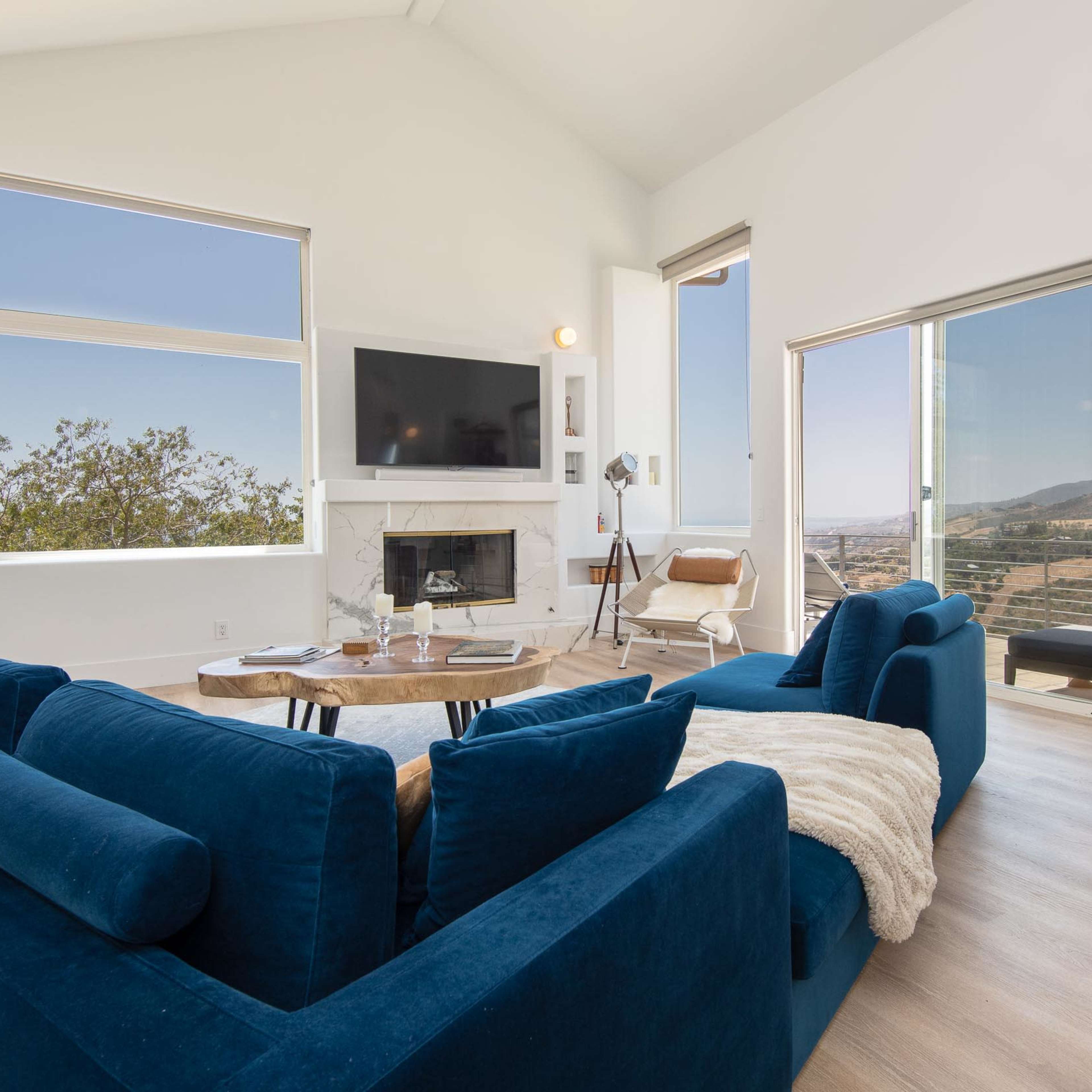 A modern living room with large windows, a fireplace, a blue sofa, and light wooden flooring.