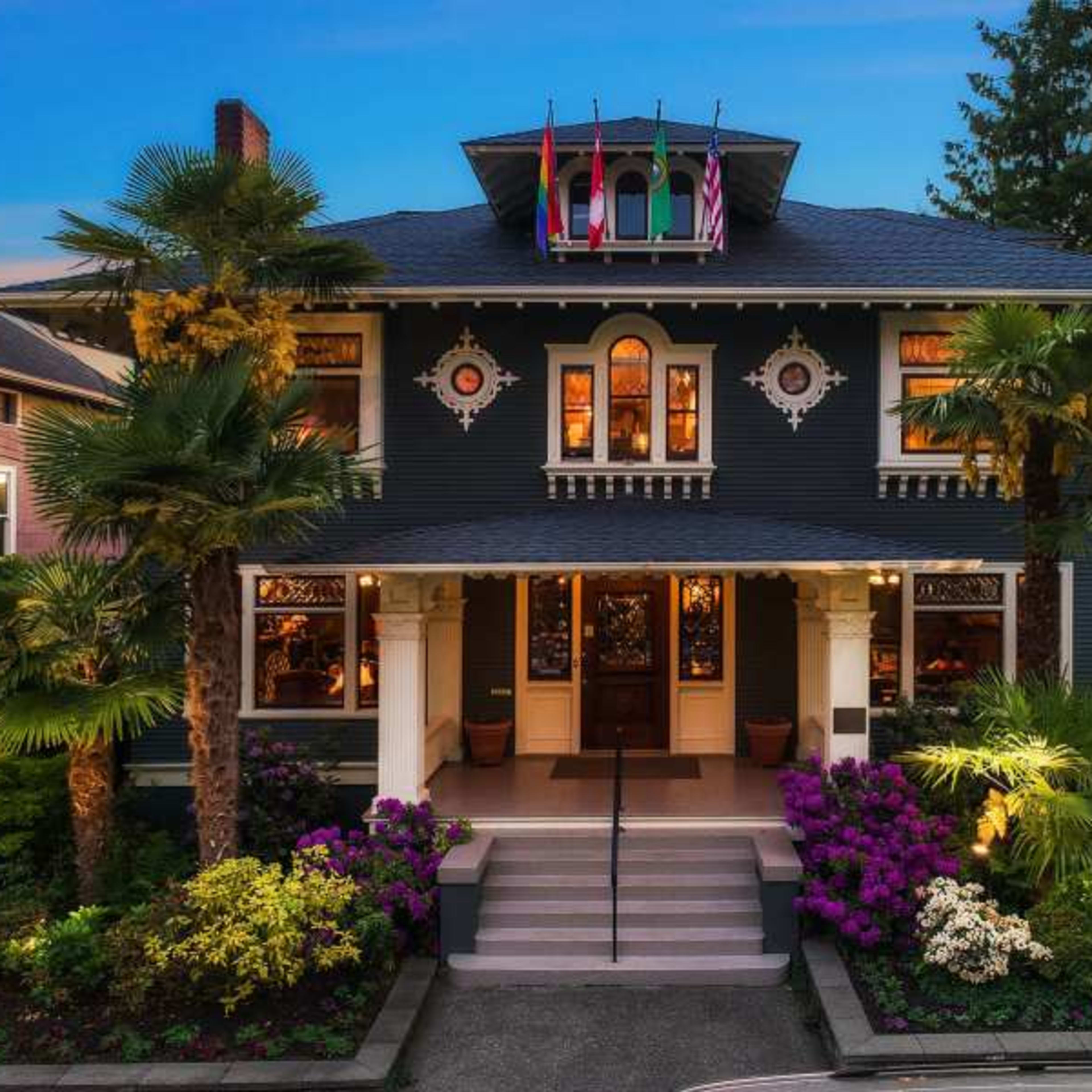 A large, two-story house adorned with palm trees and colorful flower beds, featuring decorative architectural elements and multiple flags displayed on the roof.