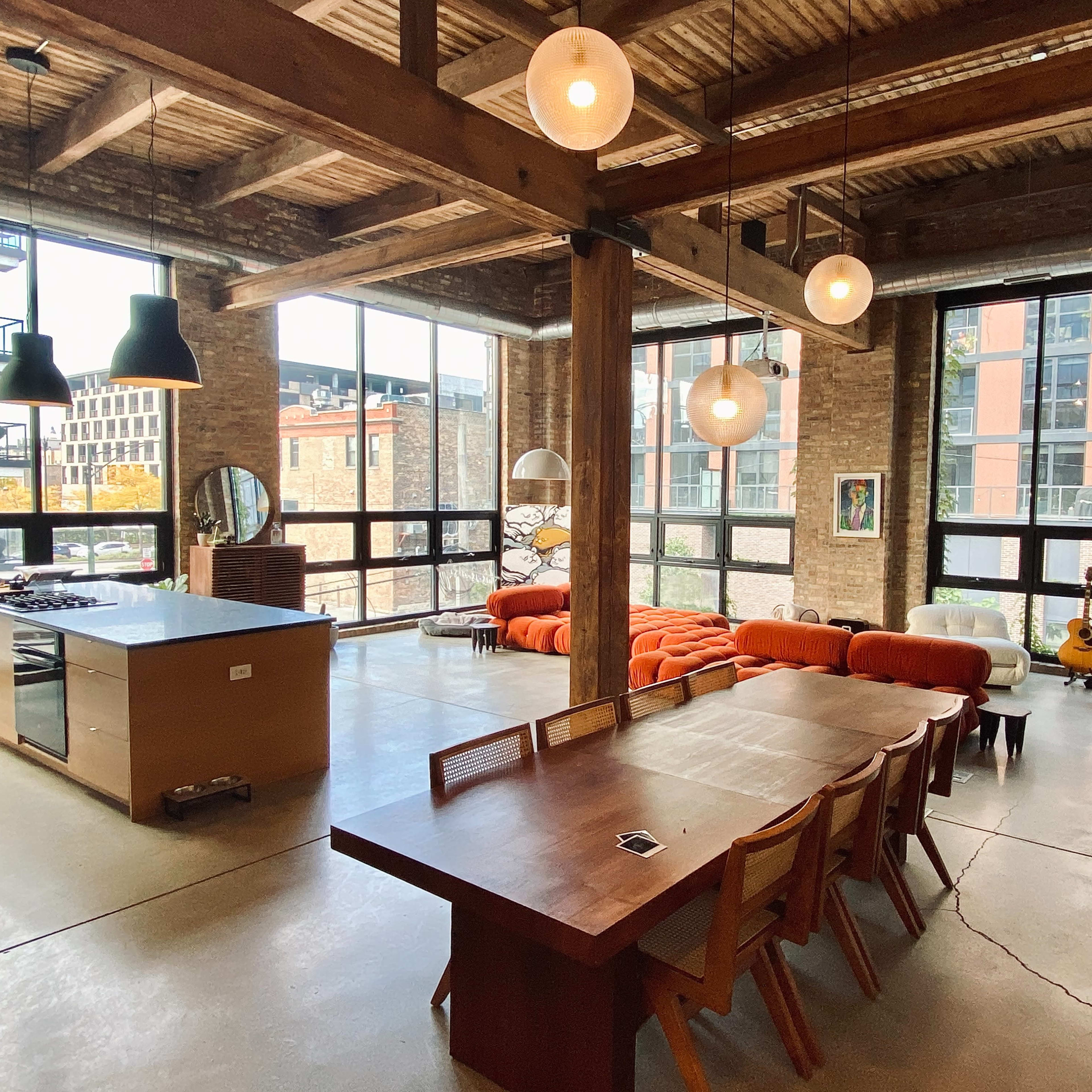A modern, open-concept living space with a kitchen, dining area, and seating area featuring a long wooden table, orange couch, and large windows allowing natural light.