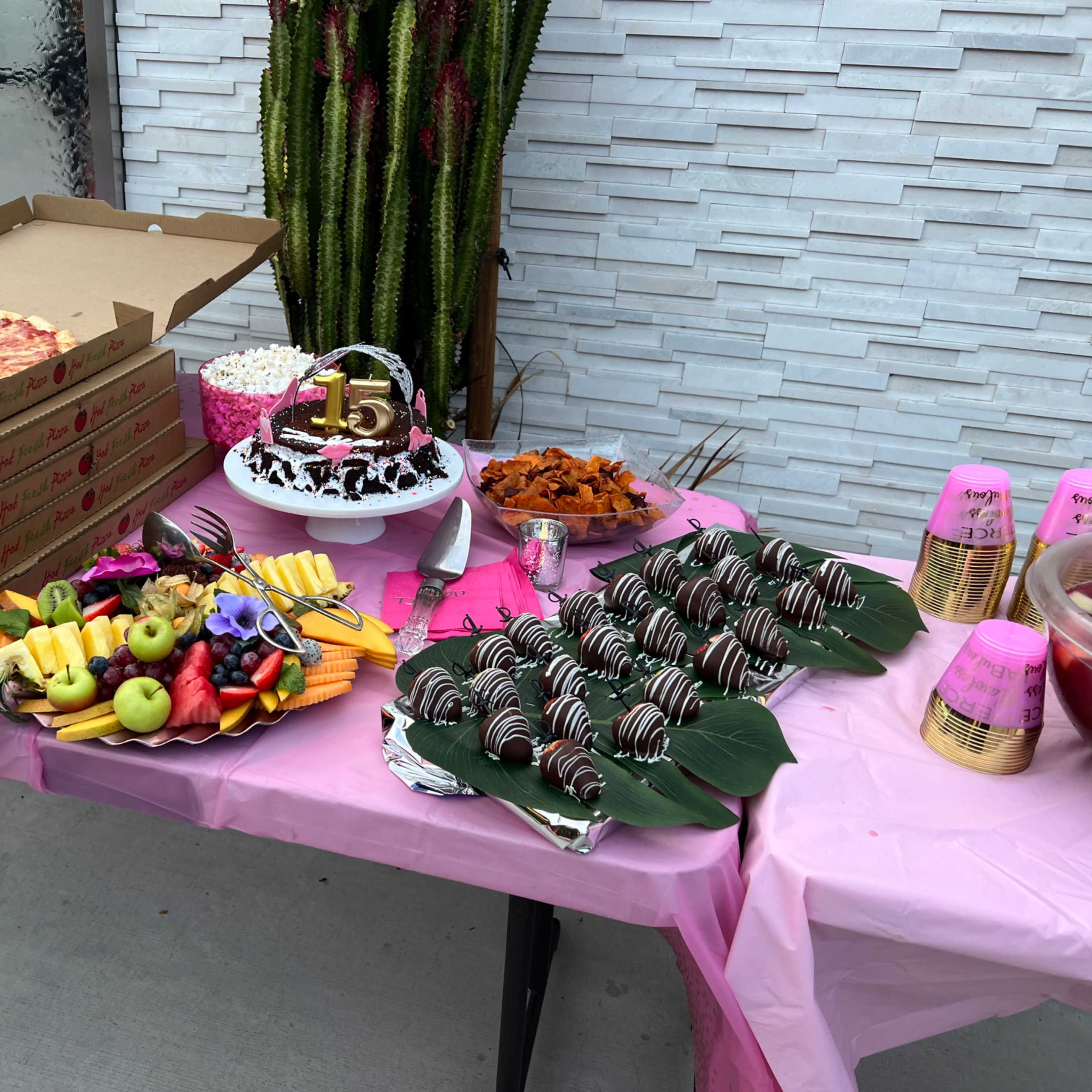 A table displays a variety of food items, including pizza boxes, a chocolate cake topped with the number 15, fruit arrangements, strawberries dipped in chocolate, and cups.